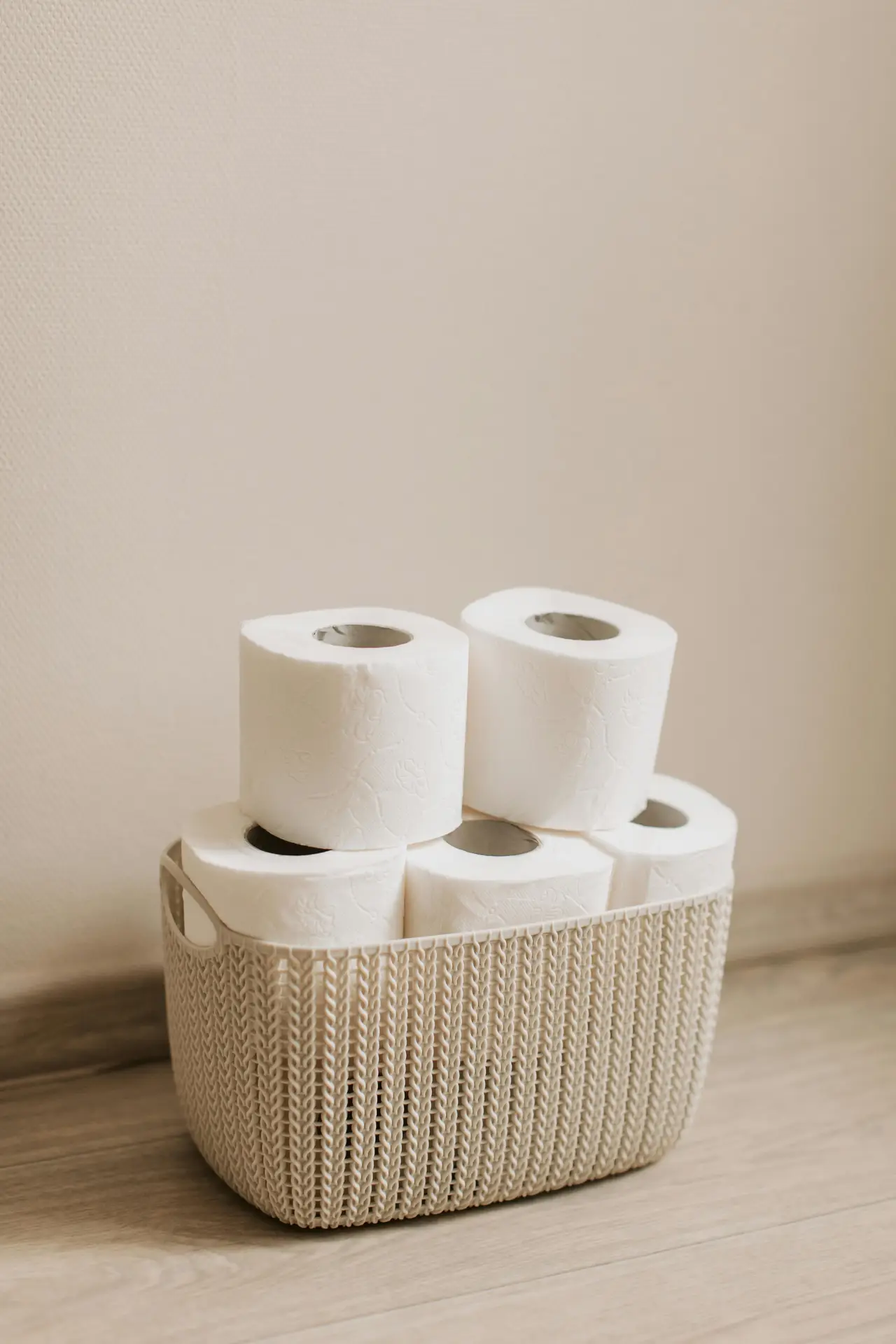 A woven-look toilet paper bin placed on a wooden floor holds several neatly stacked white rolls against a soft beige wall. This simple setup highlights smart organization ideas for the bathroom, offering an easy way to keep extra supplies accessible while maintaining a clean and coordinated look.