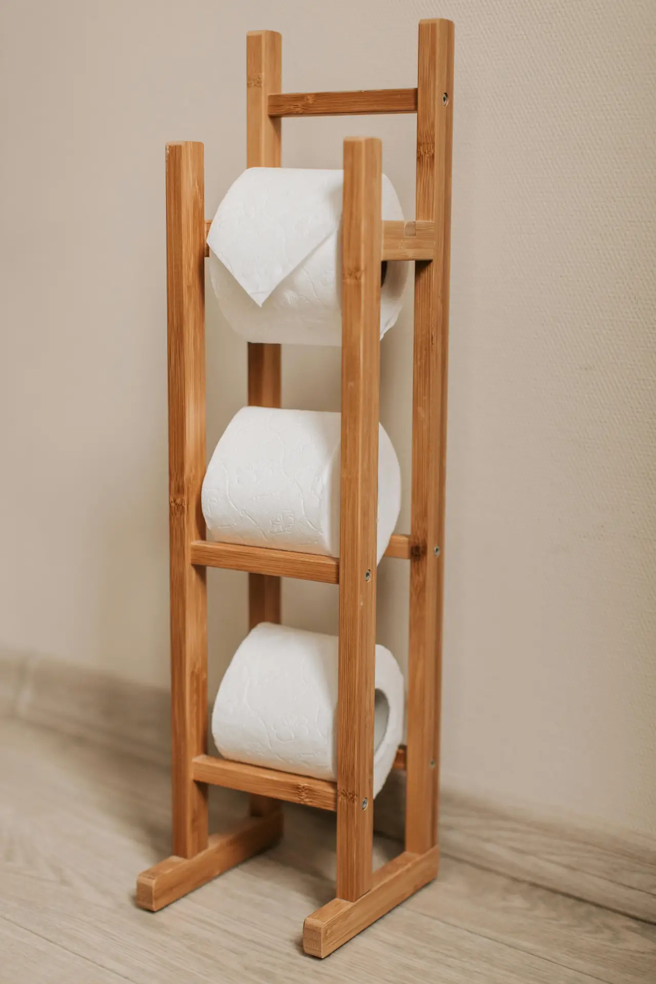 Freestanding bamboo toilet paper holder designed as a slim ladder-style stand, neatly storing three white rolls against a neutral wall and wood-look floor, offering practical organization ideas for the bathroom with a compact and stylish storage solution.
