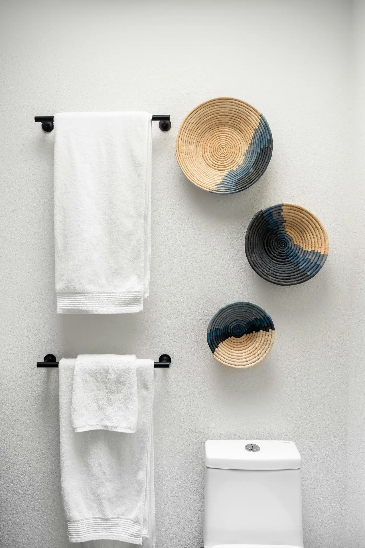 Minimalist bathroom wall featuring two black towel bars on the wall holding neatly folded white towels, paired with three woven decorative baskets in neutral and navy tones. The top of a white toilet is visible below, showcasing simple organization ideas for the bathroom that blend function with subtle wall decor.