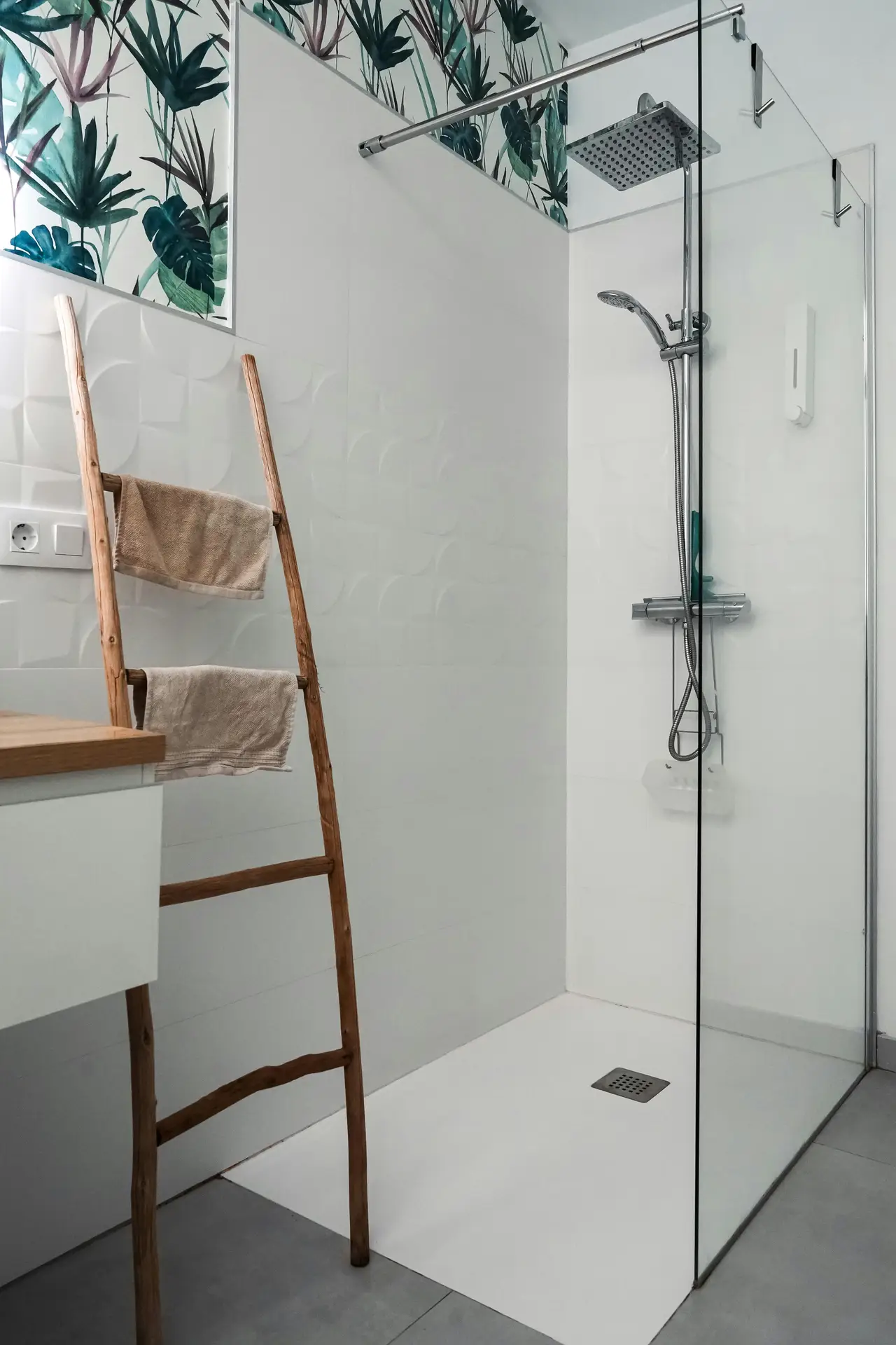 Bright, modern bathroom with a walk-in glass shower, rainfall showerhead, and white walls accented by tropical leaf wallpaper near the ceiling. A slim wooden towel ladder leans against the wall holding neutral-toned towels beside a floating vanity-an inspiring example of organization ideas for the bathroom that combines style and function with a space-saving towel ladder.