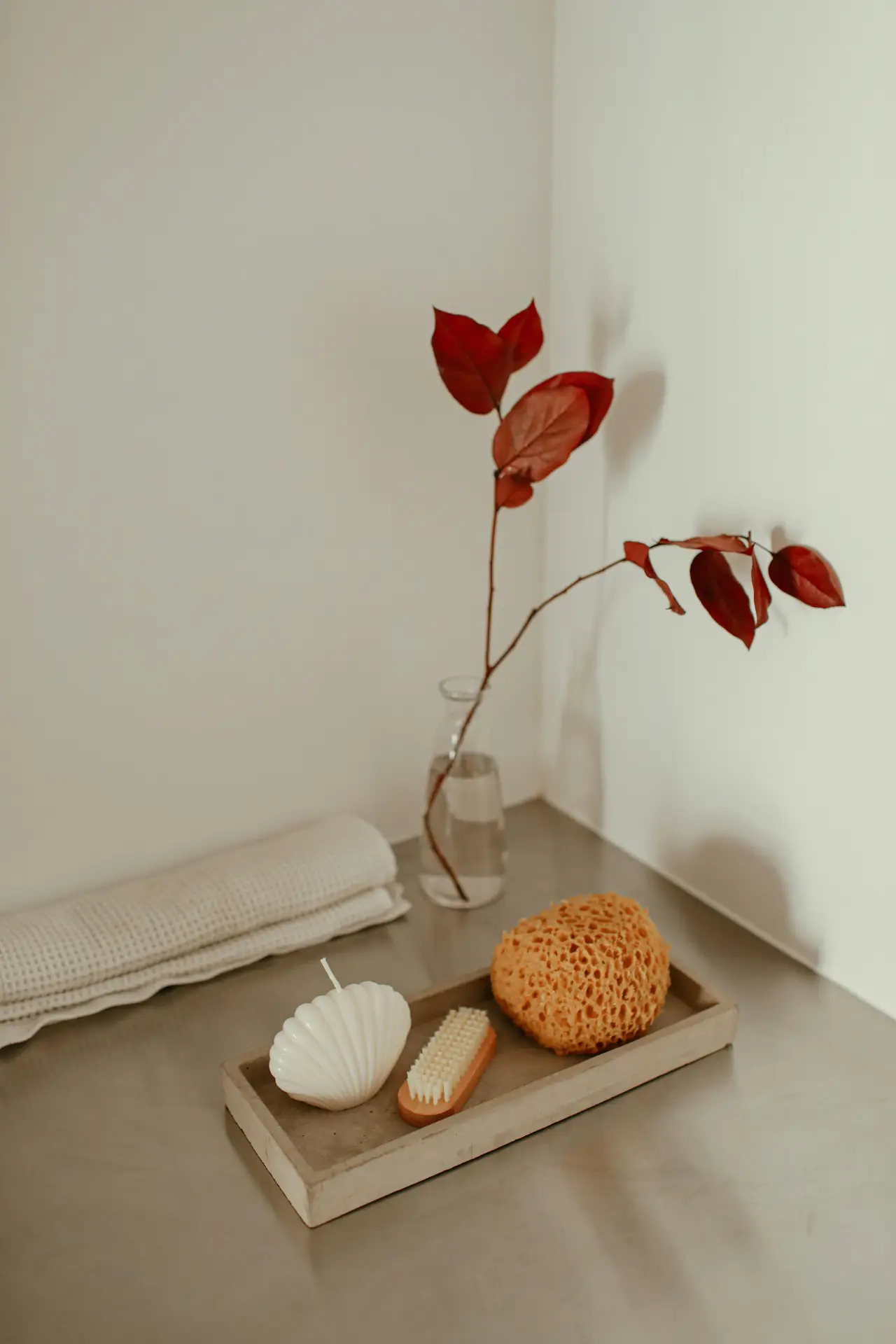 Soft, minimalist bathroom corner styled with a small wooden tray holding a seashell-shaped candle, a natural nail brush, and a sponge, while a folded towel and a glass vase with red leaves add warmth to the neutral setting. This serene arrangement highlights organization ideas for the bathroom using trays to keep everyday essentials neatly grouped and visually appealing.