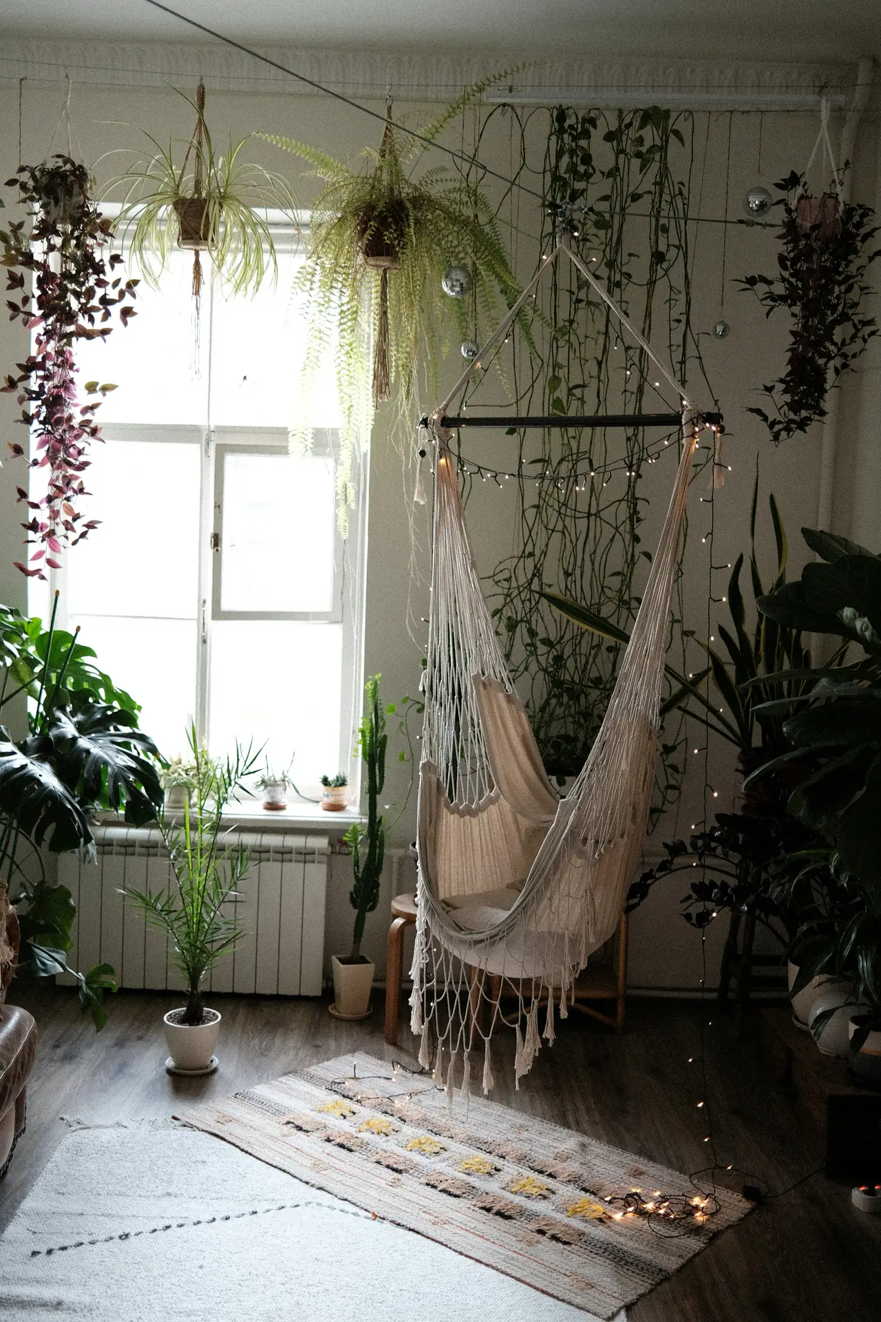 Boho-inspired corner featuring an indoor hammock suspended near a bright window, showcasing creative small living room ideas apartment residents can try. The space is filled with hanging plants, trailing vines, and potted greenery, while soft string lights and layered rugs add warmth. Natural textures, wood flooring, and abundant foliage transform a compact area into a relaxed retreat perfect for reading or unwinding.
