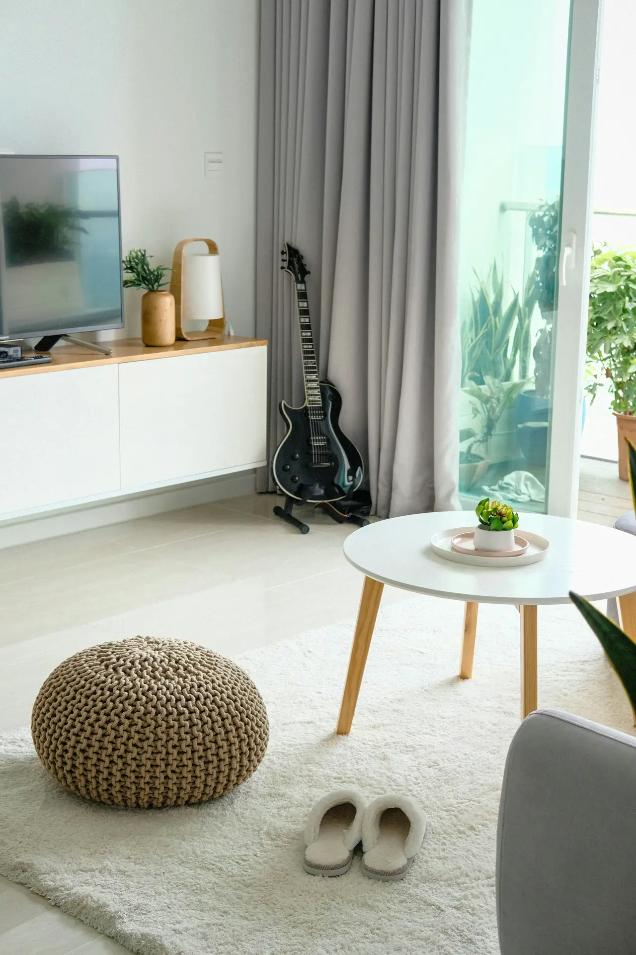 Airy, modern setup illustrating small living room ideas apartment dwellers can try, designed to use light colors for a spacious feel. A white media console, pale walls, soft gray curtains, and a light area rug brighten the room, while a round coffee table and knitted pouf add texture. A guitar by the window, indoor plants, and simple decor create a calm, uncluttered atmosphere that maximizes natural light and openness.