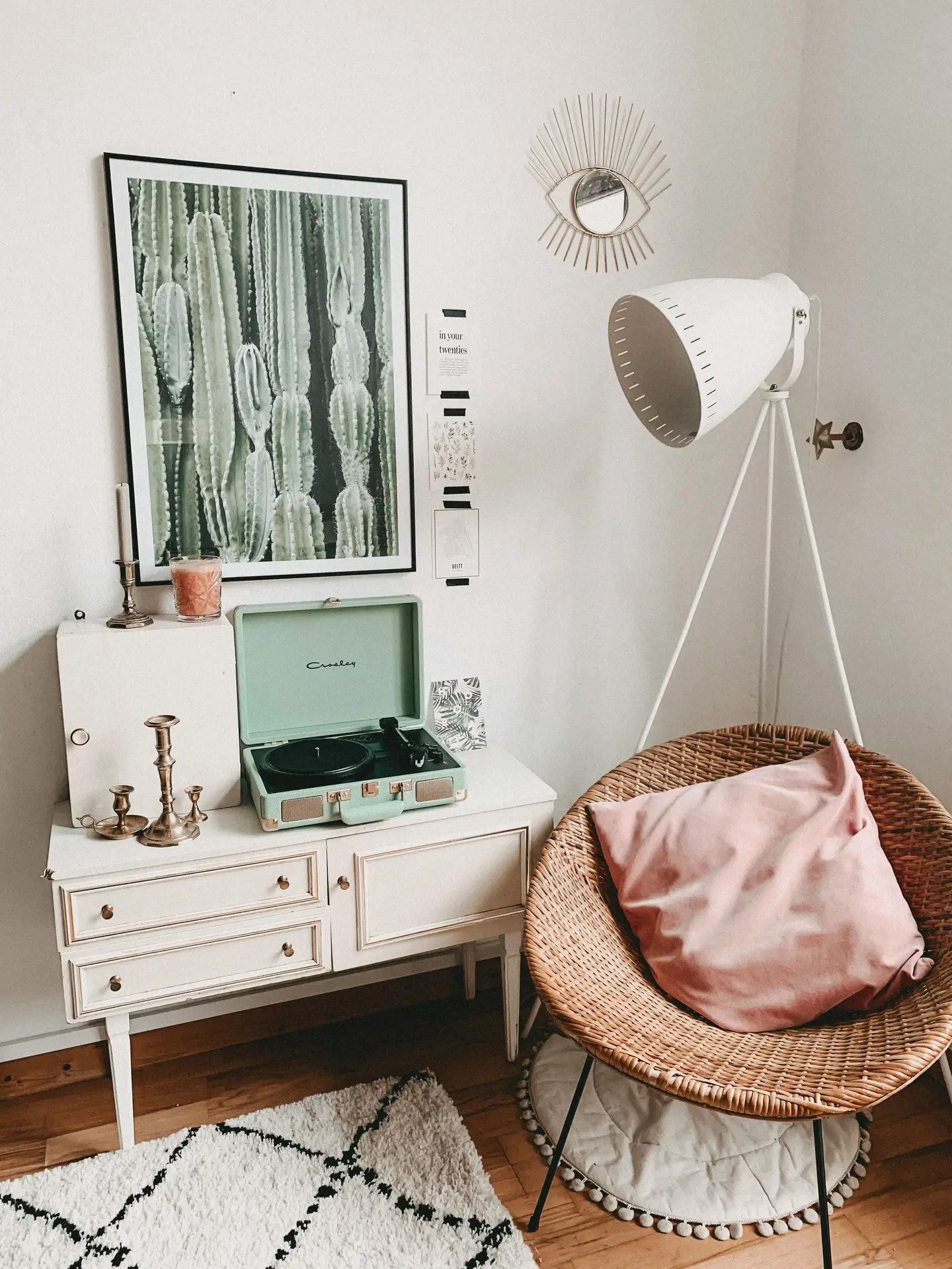 Charming corner styled as a small music niche, illustrating small living room ideas apartment owners can easily recreate. A white vintage-style sideboard holds a mint green record player beneath cactus artwork, surrounded by brass candlesticks and soft decor accents. A woven rattan chair with a blush cushion and a tall tripod floor lamp complete the cozy setup, turning an unused wall into a functional and inviting listening space.