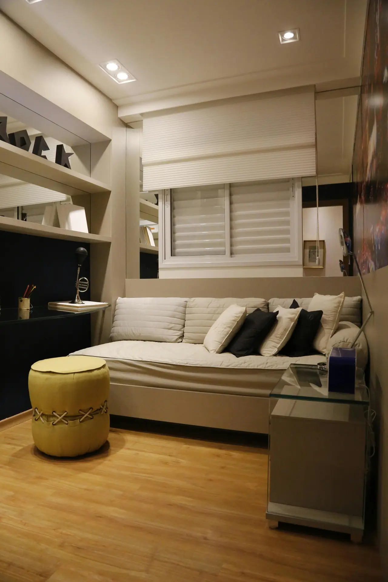 Compact lounge setup highlighting small living room ideas apartment owners can use, featuring niche fitting furniture tailored to the room's layout. A built-in sofa fits neatly beneath the window, surrounded by custom shelving and cabinetry that maximize storage. Warm wood flooring, layered cushions, a small ottoman, and a slim glass side table create a functional yet comfortable space that makes the most of limited square footage.