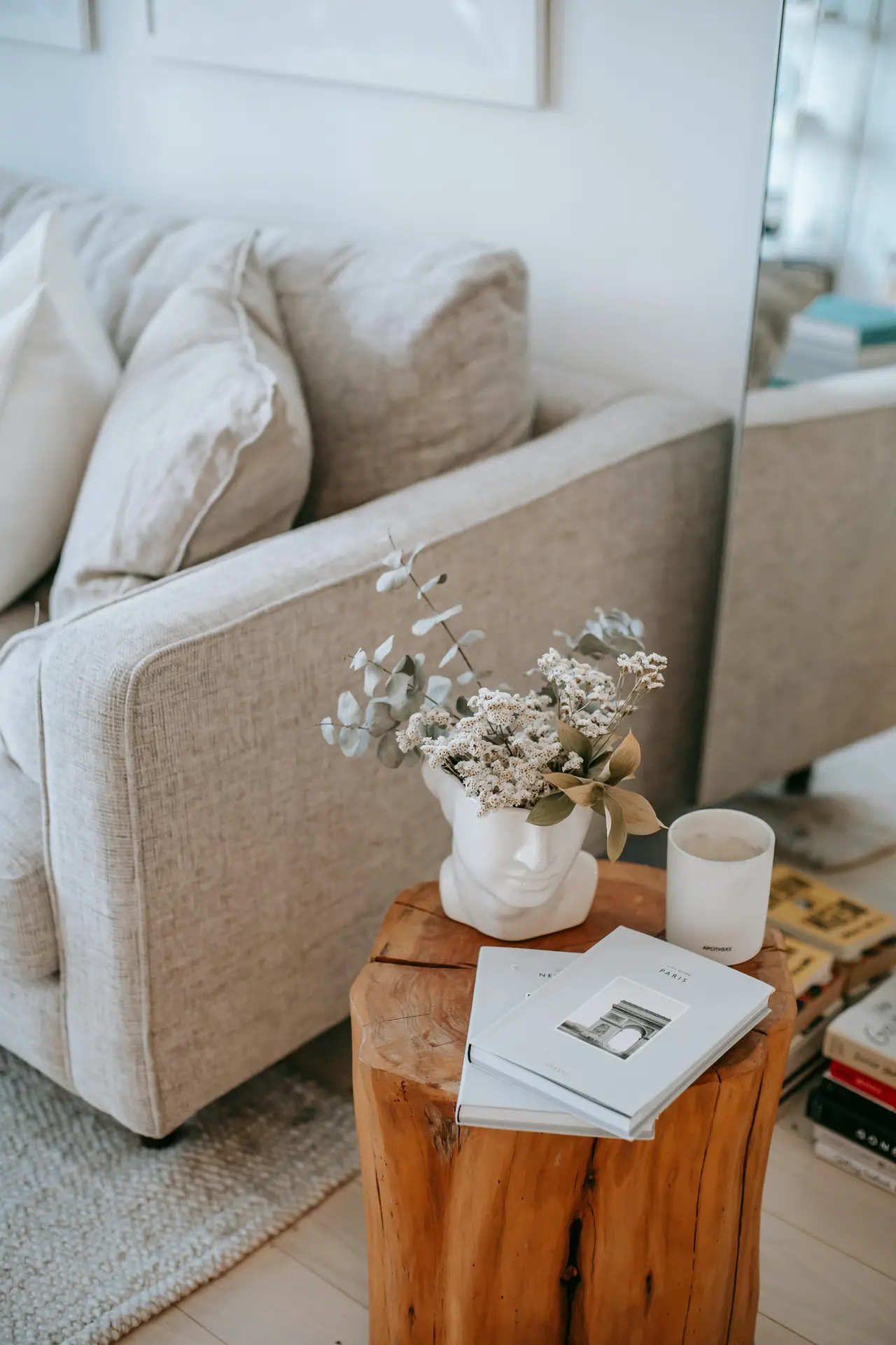 Close-up of a cozy seating corner illustrating small living room ideas apartment dwellers can recreate, featuring one of the trending small side tables made from a natural wood stump. The table holds stacked design books, a minimalist candle, and a sculptural face vase filled with dried flowers. A neutral fabric sofa, soft cushions, woven rug, and a leaning mirror in the background enhance the warm, compact layout.