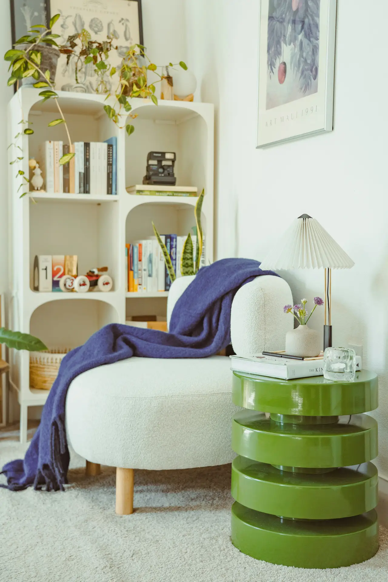 Cozy corner styled with small living room ideas apartment dwellers can easily replicate, featuring a soft upholstered lounge chair draped with a deep blue throw. A glossy green stacked side table adds a bold splash of color beside a pleated table lamp, books, and a small vase of flowers. In the background, an open shelving unit with plants, books, and decorative objects creates a fresh, inviting atmosphere perfect for compact spaces.