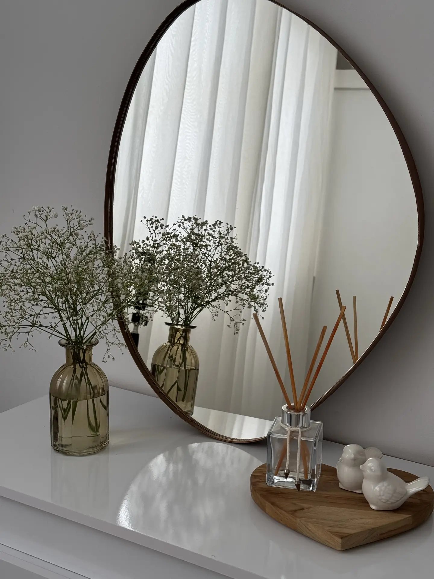 Minimal entryway styling inspired by 12 Stunning Minimalist Decor Ideas For Spring, showing a round wall mirror above a glossy white surface, with a glass vase filled with Baby's Breath, a simple reed diffuser on a wooden tray, and small ceramic bird figures, softly reflected in natural daylight through sheer curtains.