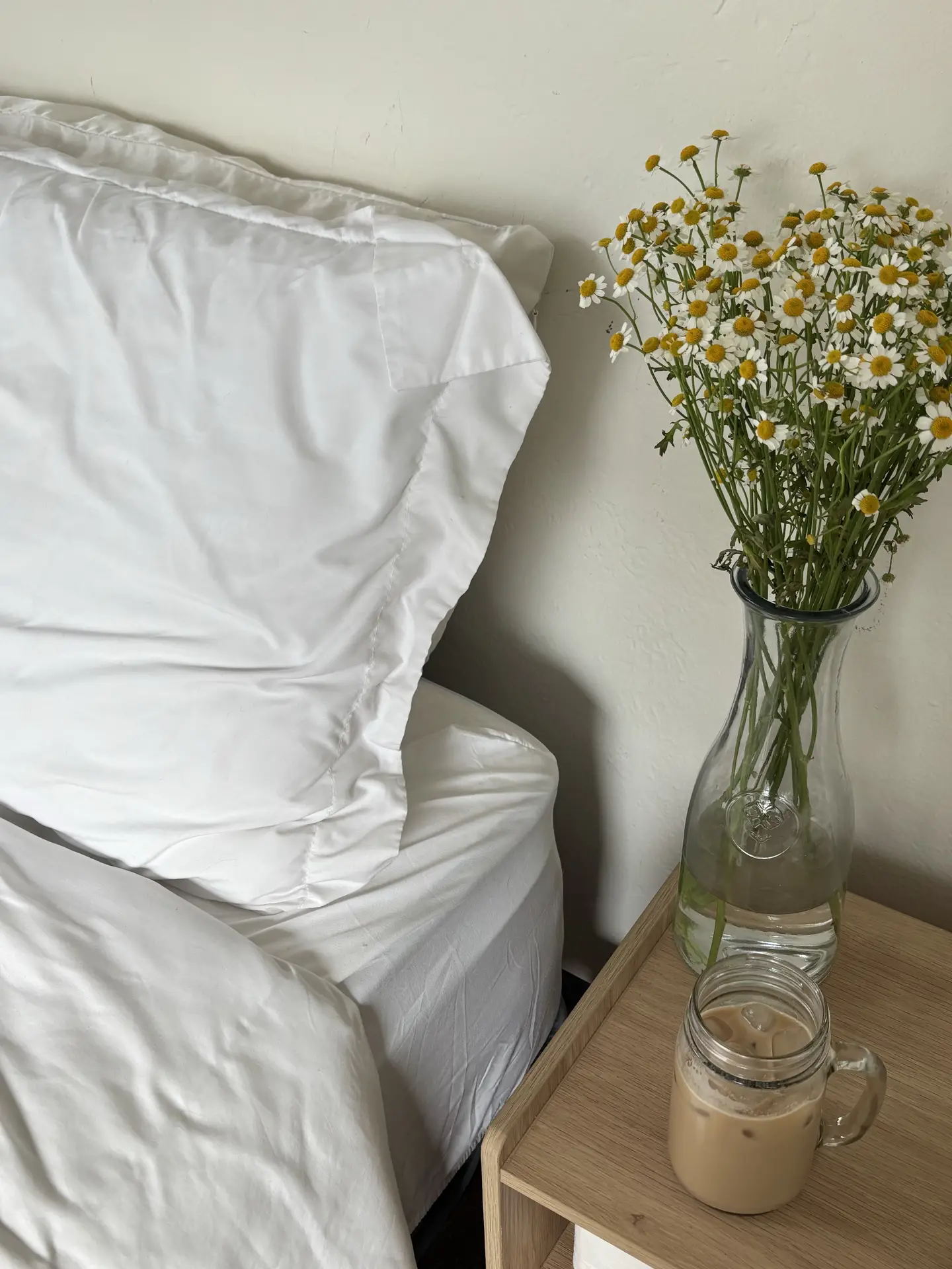 A calm bedroom corner with crisp white bedding next to a light wooden bedside table, styled with a clear glass vase filled with fresh daisies and a small jar of iced coffee. The soft colors and simple textures reflect 12 Stunning Minimalist Decor Ideas For Spring, showing how daisies can bring a relaxed, airy touch to everyday home styling.