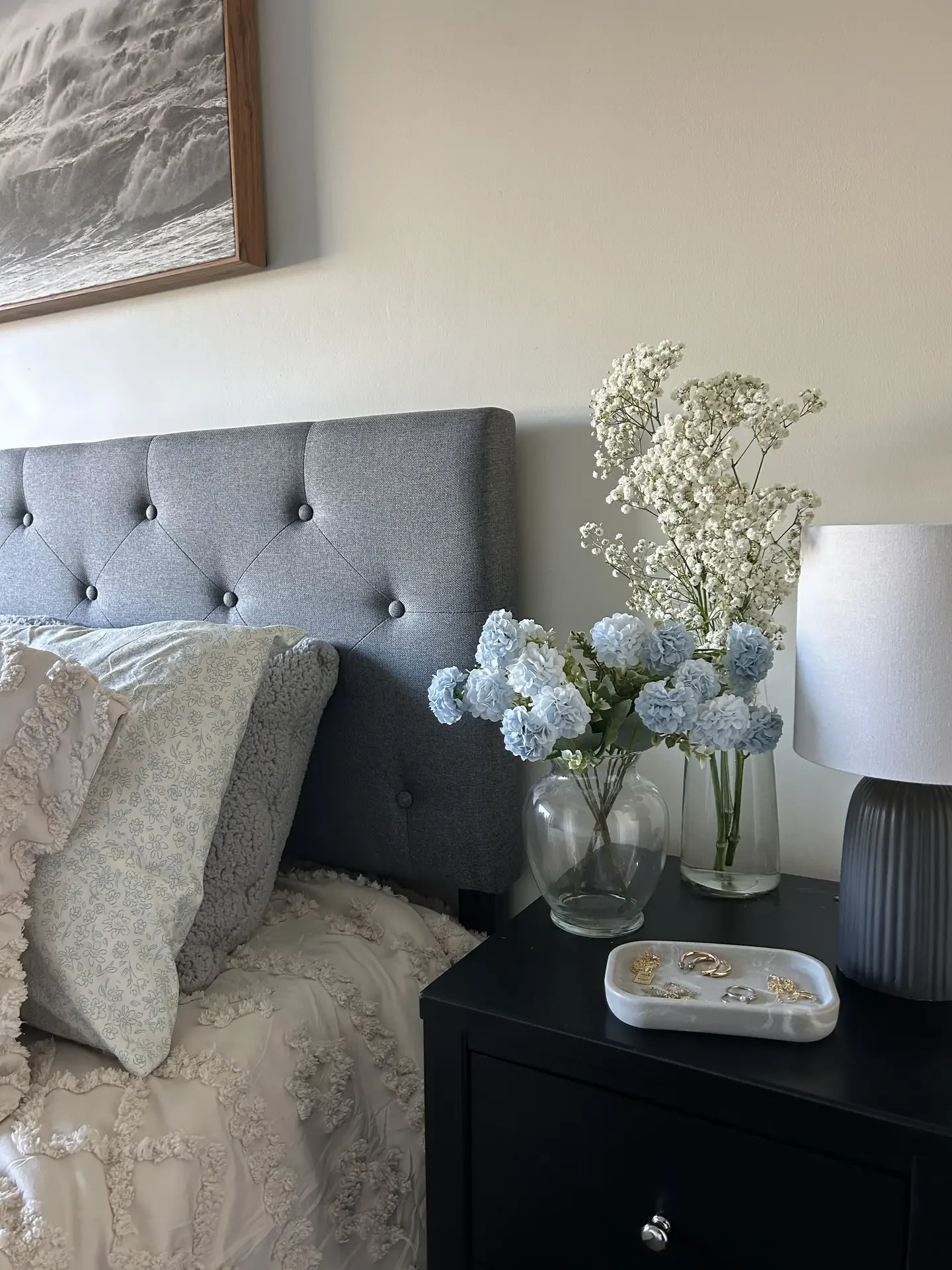 Calm bedroom styling inspired by 12 Stunning Minimalist Decor Ideas For Spring, showing light blue hydrangeas & baby's breath arranged in clear glass vases on a dark bedside table, paired with soft layered bedding, textured pillows, and a grey upholstered headboard for an airy, serene spring atmosphere.