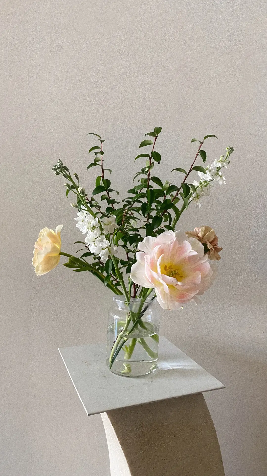 Minimalist floral arrangement displayed on a small sculptural pedestal, featuring light pink, yellow & white flowers mixed with fresh green stems in a clear glass vase. The soft color palette and simple styling create an airy, modern look that fits perfectly with 12 Stunning Minimalist Decor Ideas For Spring, highlighting how seasonal flowers can add subtle elegance to a calm interior setting.