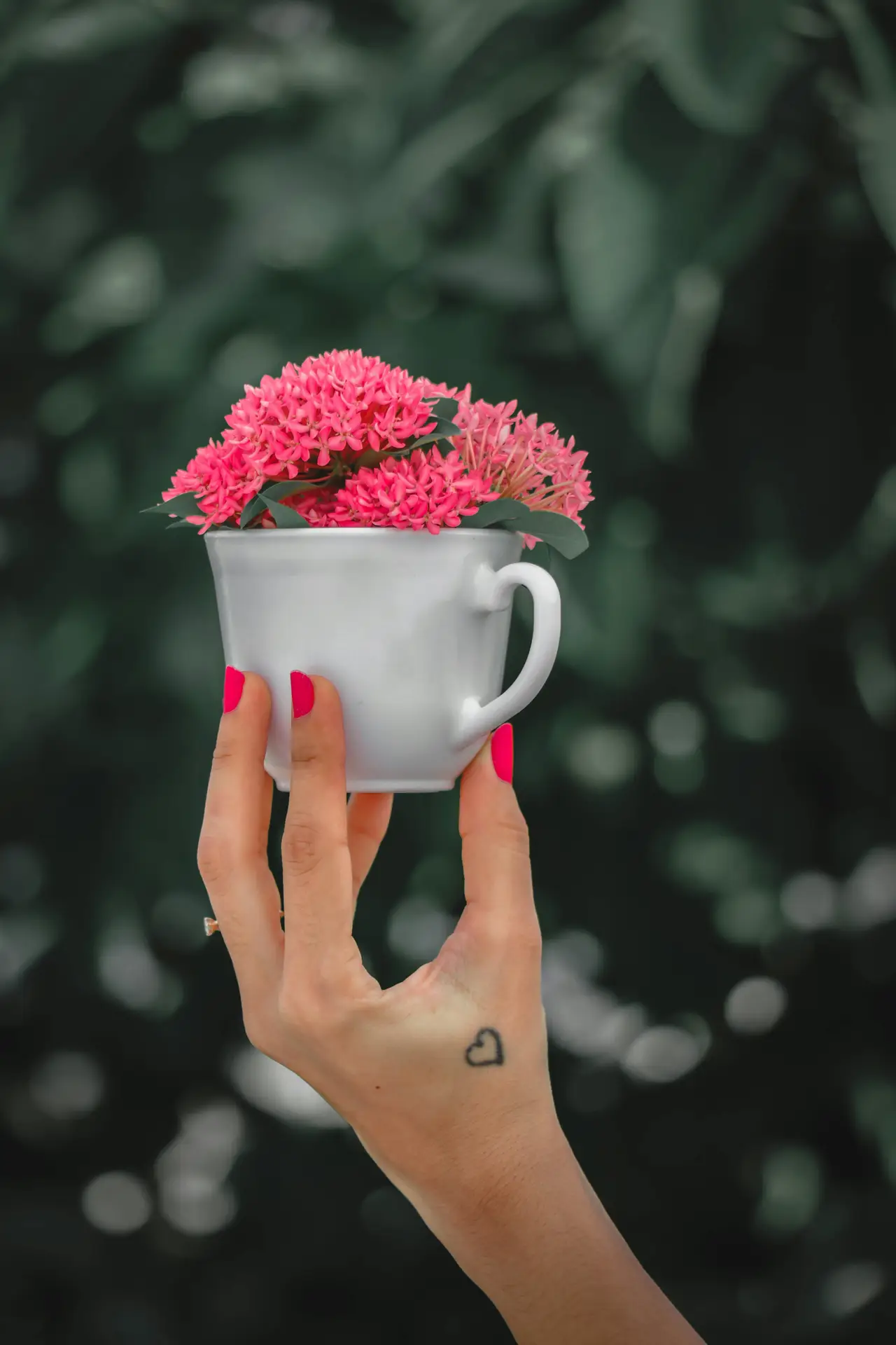 Creative spring detail inspired by 12 Stunning Minimalist Decor Ideas For Spring, featuring a hand holding a white ceramic cup filled with pink ixora in a cup, set against a softly blurred green background, with bright nail polish and a small heart tattoo adding a personal, modern touch.
