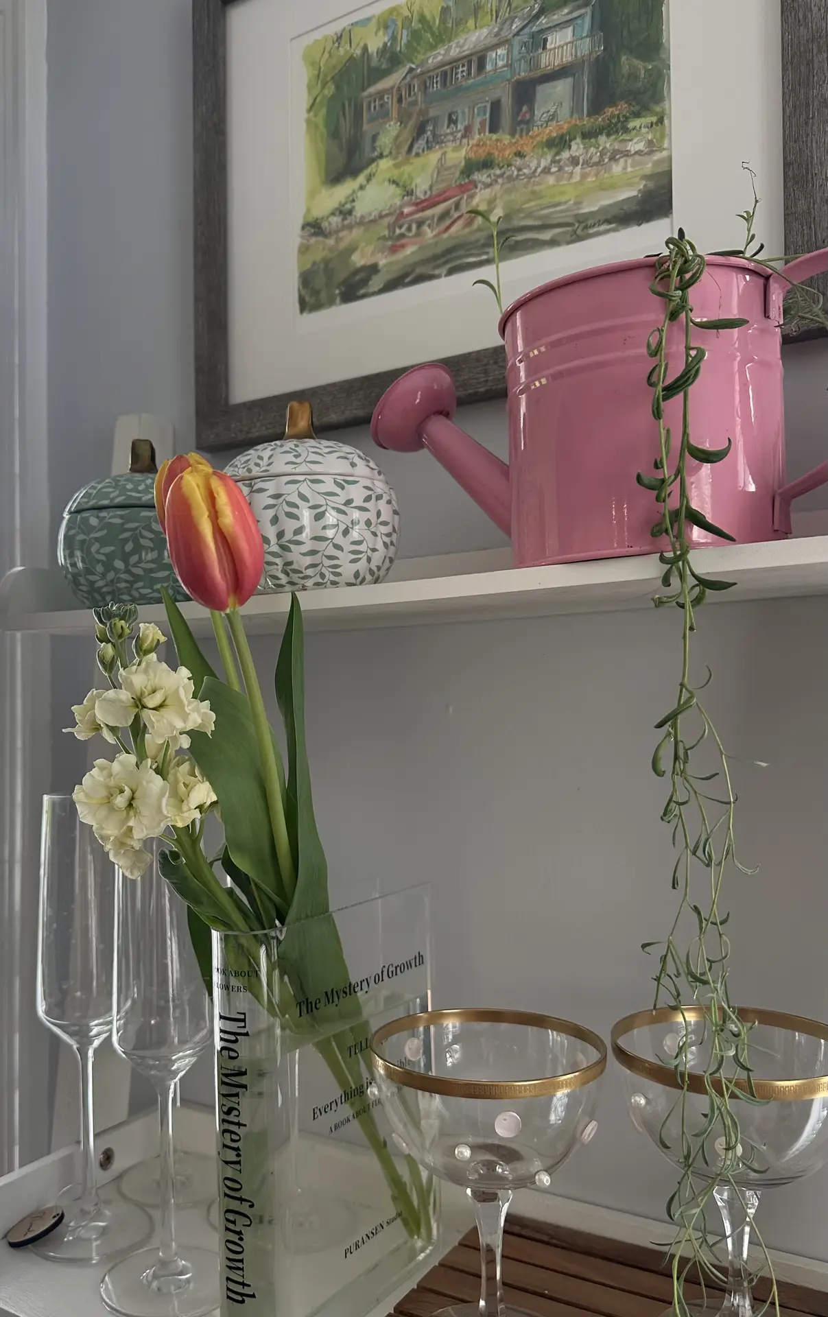 Fresh shelving vignette inspired by 12 Stunning Minimalist Decor Ideas For Spring, featuring a pink watering can and spring flowers arranged with glass vases, tulips, and delicate white blooms, complemented by decorative bowls, hanging greenery, and a framed artwork for a light and curated home styling moment.