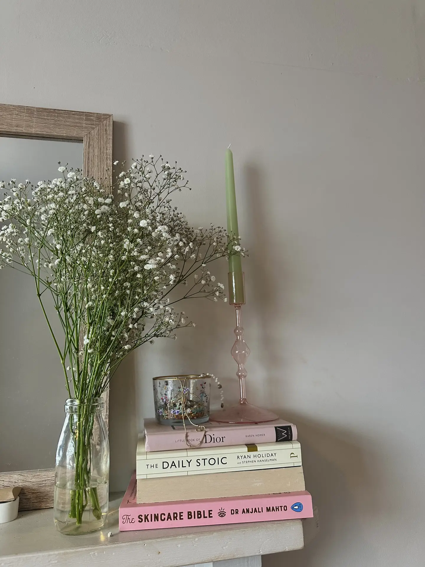 Soft spring vignette inspired by 12 Stunning Minimalist Decor Ideas For Spring, showing stacks of books in pastel colors topped with a delicate glass candle holder and small decorative jar, next to a simple vase filled with airy white flowers, all set against a neutral wall with a wooden-framed mirror for a calm, minimalist look.