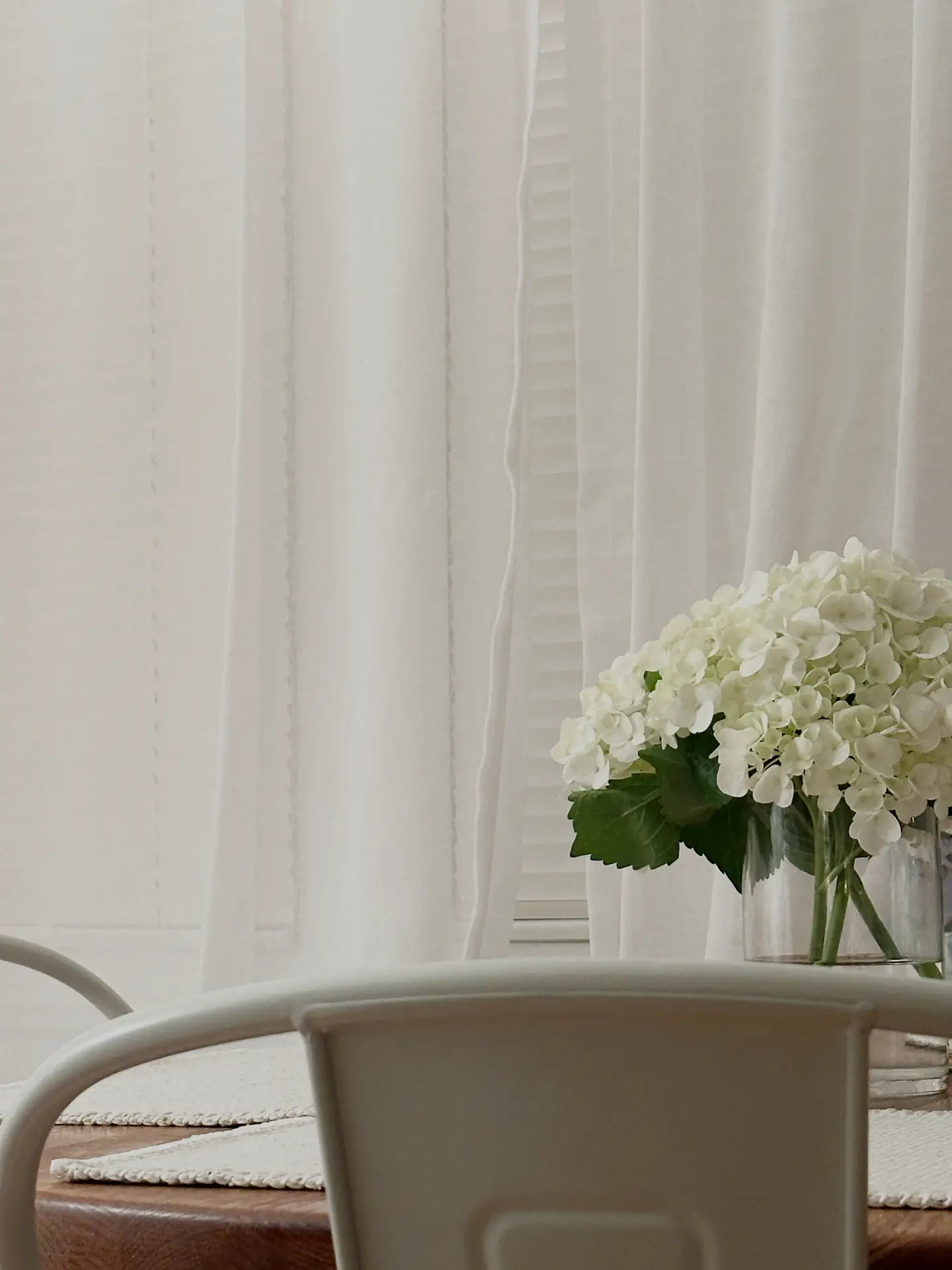 A soft, light-filled dining or seating area with sheer white curtains, a neutral chair in the foreground, and a clear glass vase holding white hydrangeas on a wooden table. The calm tones and natural light reflect 12 Stunning Minimalist Decor Ideas For Spring, showing how white hydrangeas add an elegant, fresh accent to a simple interior setting.