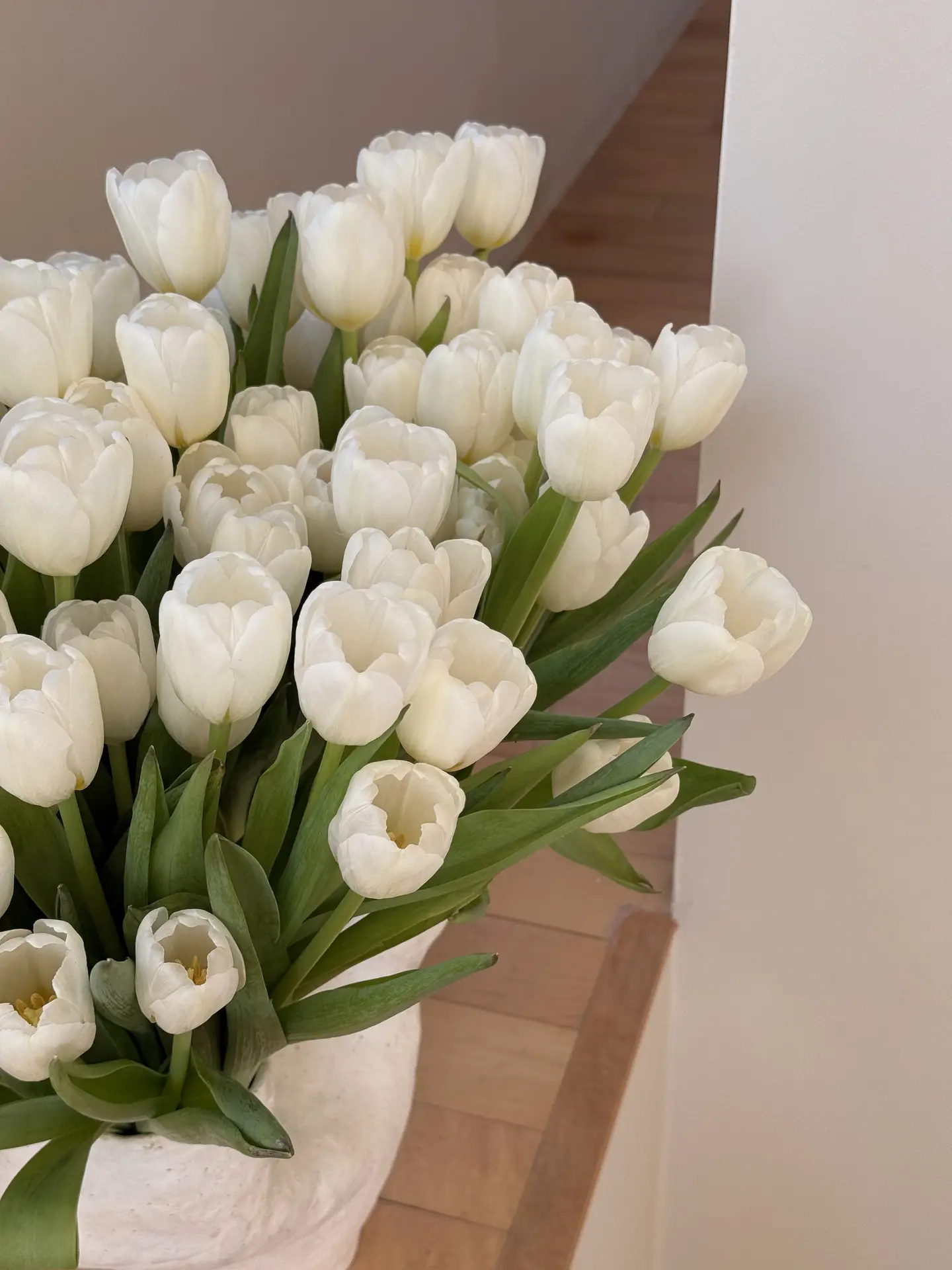 A full bouquet of white tulips arranged in a simple white vase, placed on a light wooden surface in a bright, calm interior. The soft tones and clean styling reflect the aesthetic of 12 Stunning Minimalist Decor Ideas For Spring, showing how white tulips in a white vase can create an elegant, fresh look that works in many different living spaces.