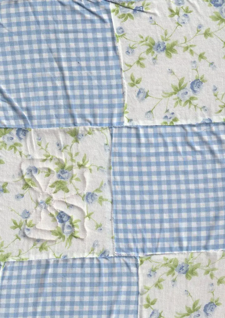 A top-down view of a light blue and white cottagecore patchwork quilt. The design alternates between a classic gingham check pattern and white fabric squares decorated with delicate blue flowers and green leaves, perfect for rustic floral bedding ideas.
