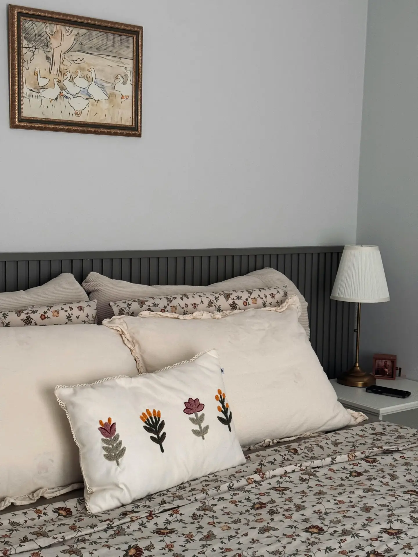 A cozy bed arrangement featuring cream-colored ruffled pillows and a white accent pillow with colorful embroidered accents of stylized flowers. The bed is layered with a floral patterned duvet, set against a dark grey slatted headboard and a bedside table with a classic lamp, providing charming floral bedding ideas.