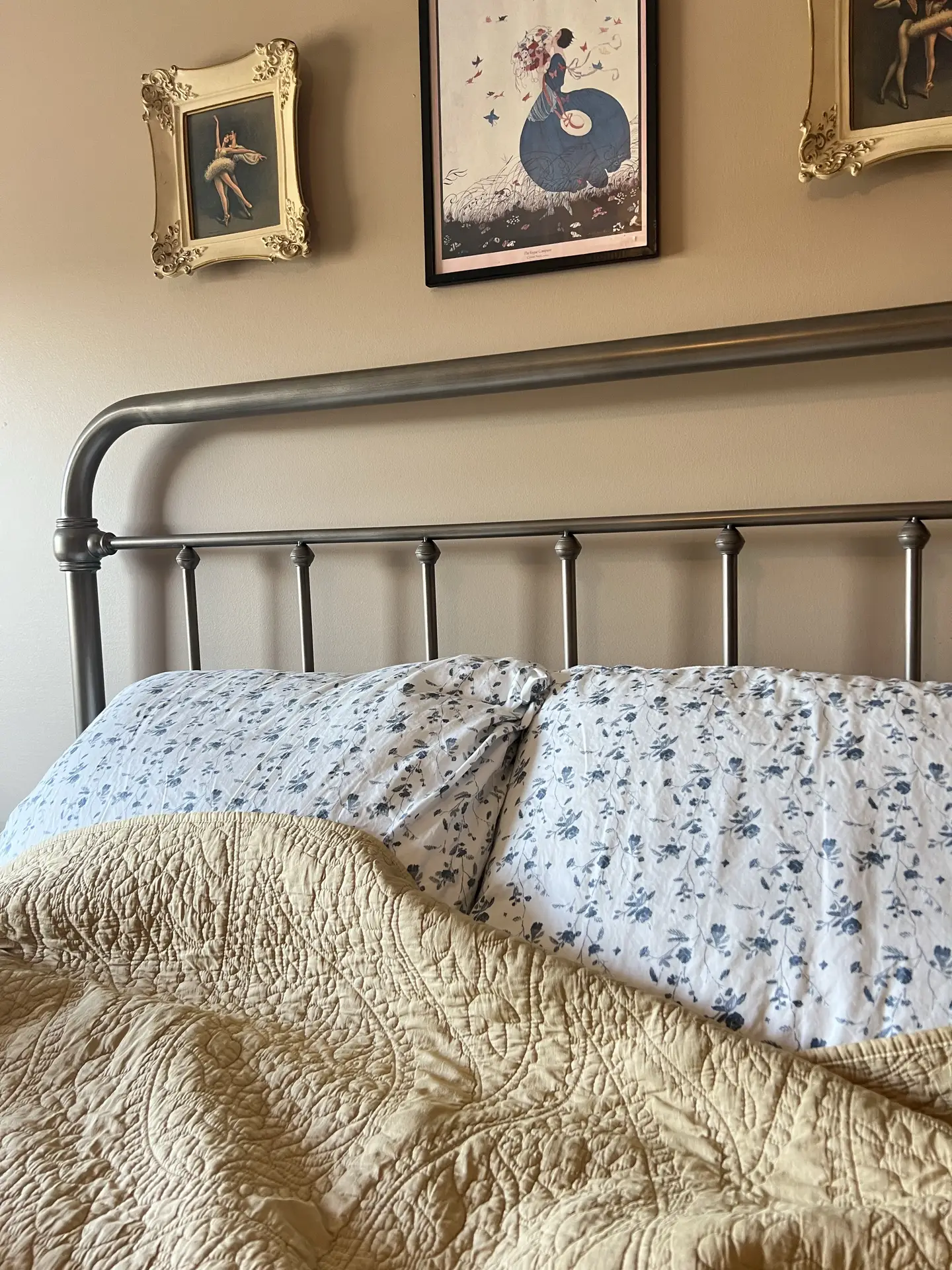 A classic metal bed frame with white pillows featuring a delicate heritage blue toil floral pattern. A quilted beige coverlet is draped over the bed, set against a tan wall decorated with vintage framed artwork, providing timeless floral bedding ideas.