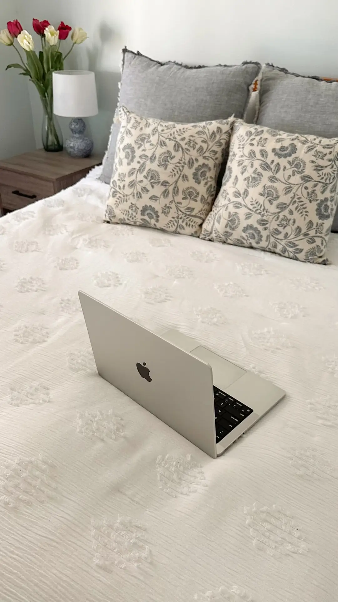 A bright, clean bedroom setting featuring a bed with a white textured duvet and two pillows with grey botanical prints. An open silver laptop sits in the foreground, while a bedside table with a lamp and a vase of red and white tulips completes the modern floral minimalist aesthetic, offering fresh floral bedding ideas.