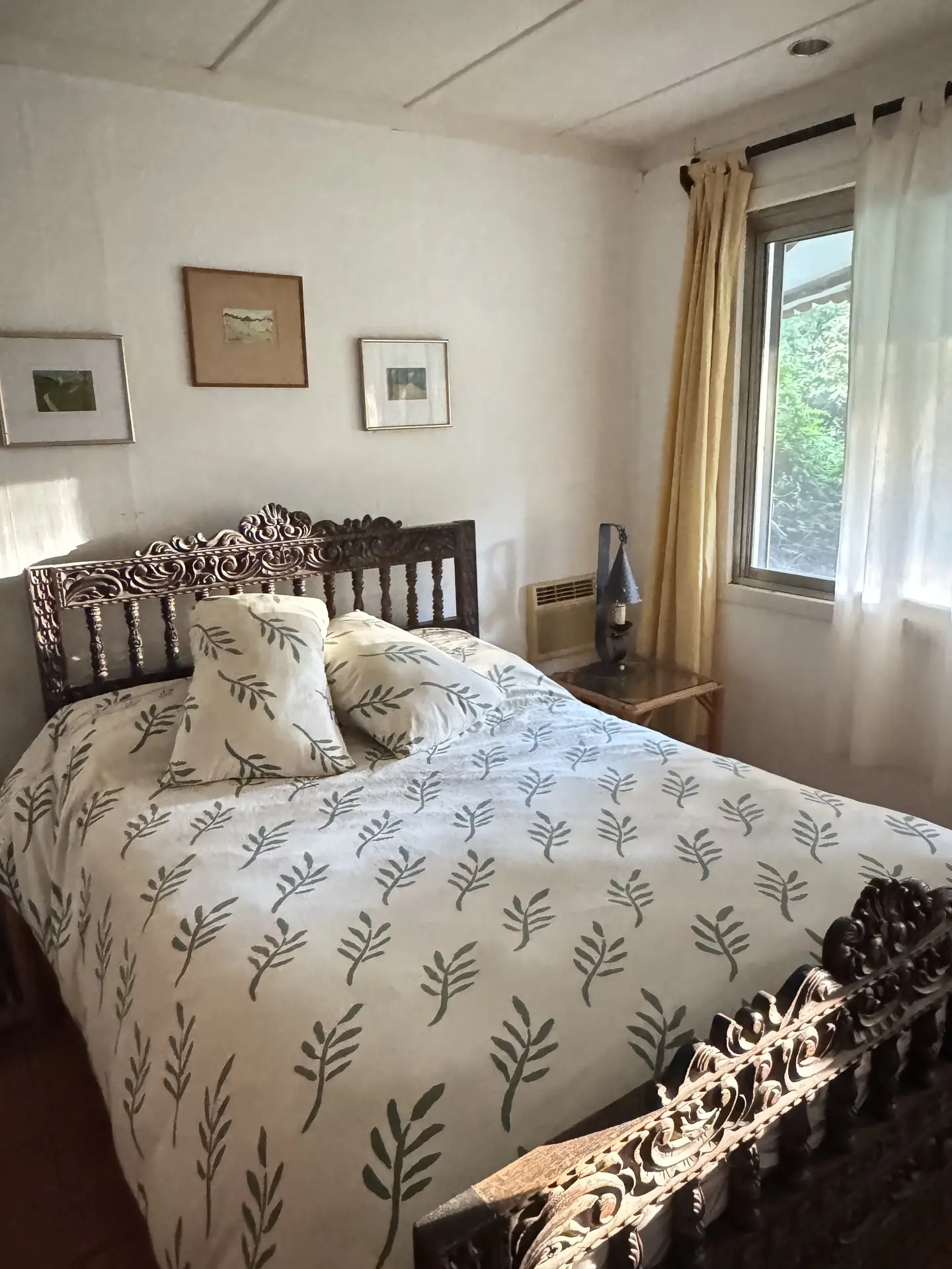 A bedroom featuring a dark, intricately carved wooden bed frame with ornate willows patterns on the white duvet and matching pillows. The sunlit room includes vintage framed art on the wall and soft yellow curtains, offering elegant floral bedding ideas for a classic interior.