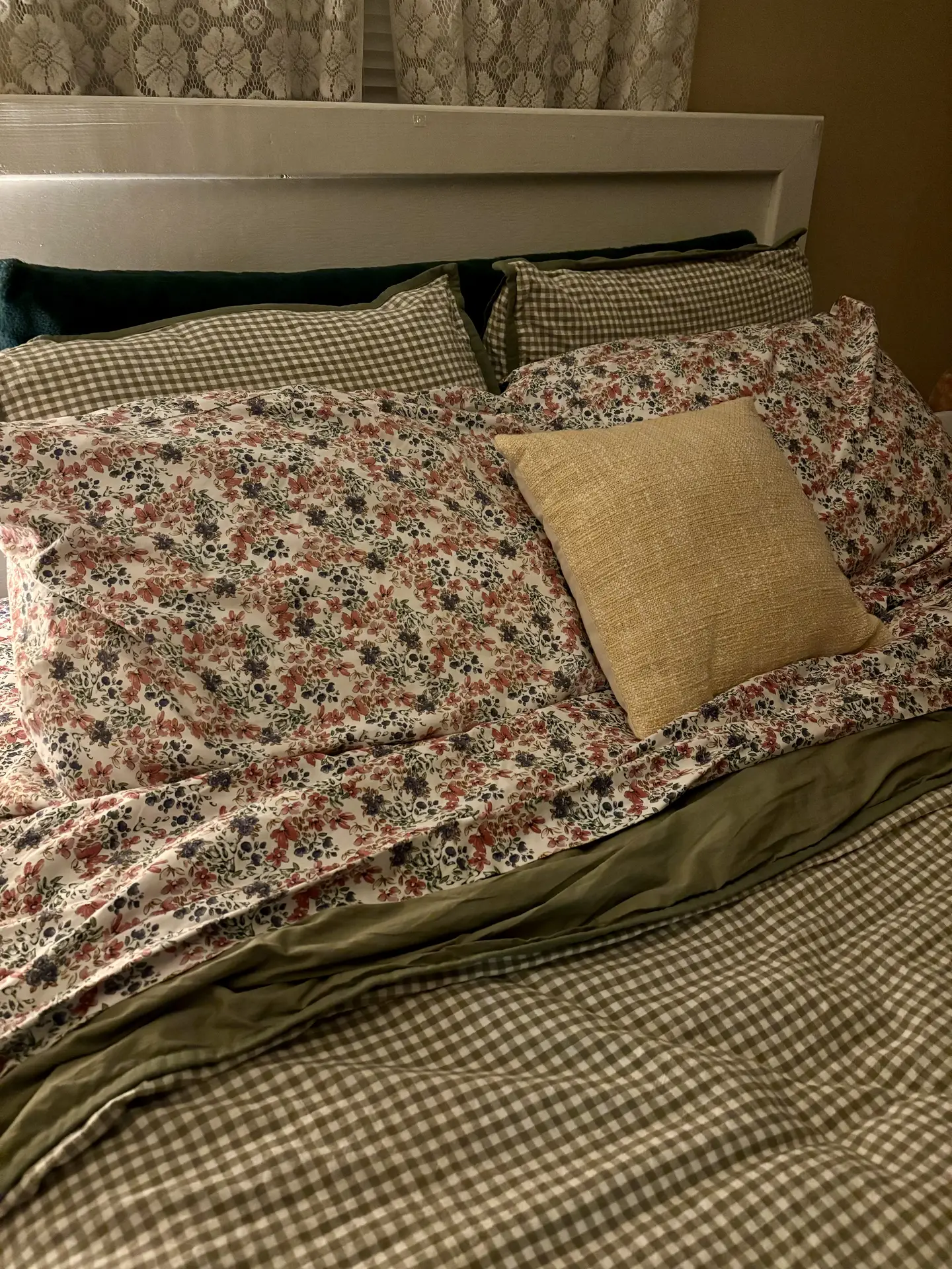 A cozy bed showcasing vintage layers with a mix of patterns, including green gingham check pillowcases and a duvet, paired with floral sheets featuring small red and purple blossoms. A single mustard-yellow textured throw pillow sits in the center, providing eclectic floral bedding ideas for a warm, lived-in look.