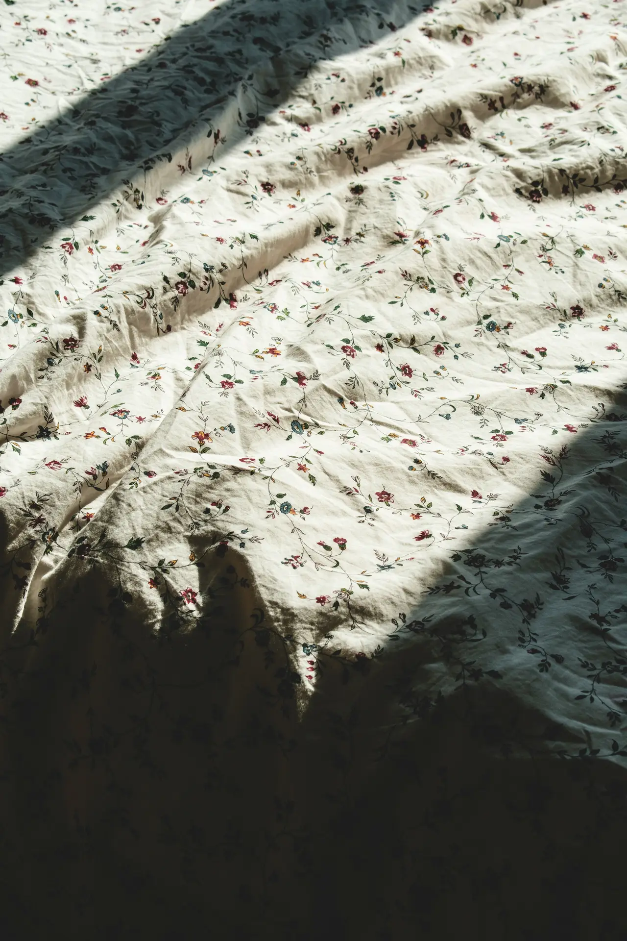 A close-up shot of rumpled cream-colored bedding featuring a delicate pattern of vintage wildflowers in shades of red, blue, and green. The soft sunlight creates a play of shadows across the fabric, offering cozy floral bedding ideas for a rustic or cottage-style bedroom.