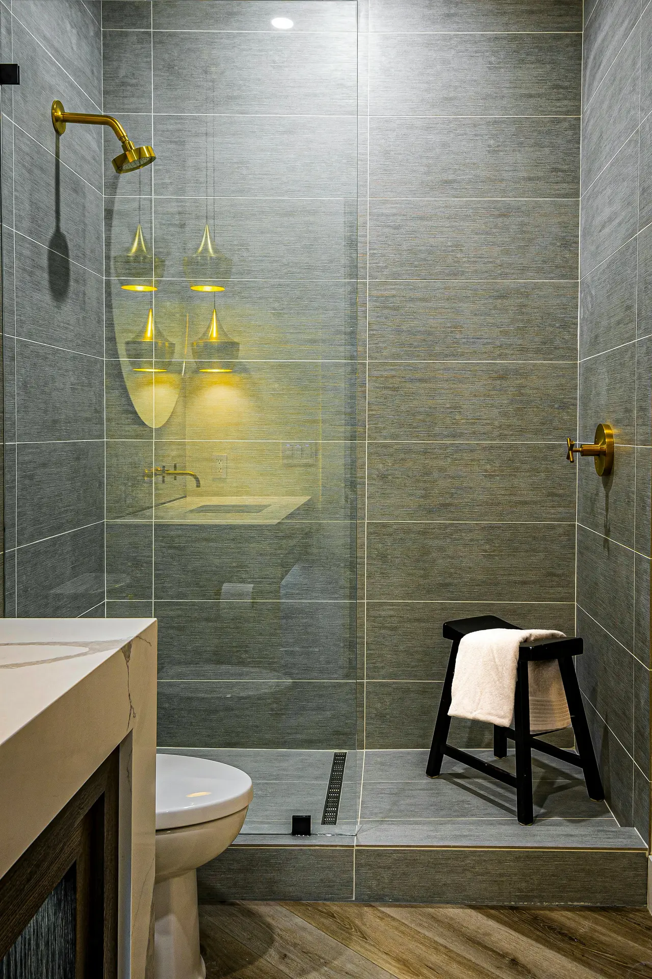 A photograph of a modern and efficiently designed guest bathroom interior. Large-format, big grey tiles clad the back feature wall and the interior of the walk-in shower, creating a sleek and durable surface. This contemporary tiling is balanced by warm white vertically slatted wainscoting on the adjacent walls. The thoughtful guest bathroom ideas are evident in the compact floating wood vanity with an integral sink and a circular mirror, paired with a modern wall-mounted toilet to save floor space. A glass shower screen divides the area, and small hexagon mosaic tiles are on the shower floor. Accents include a woven basket with rolled towels and several potted plants, adding natural textures to the cool tones.