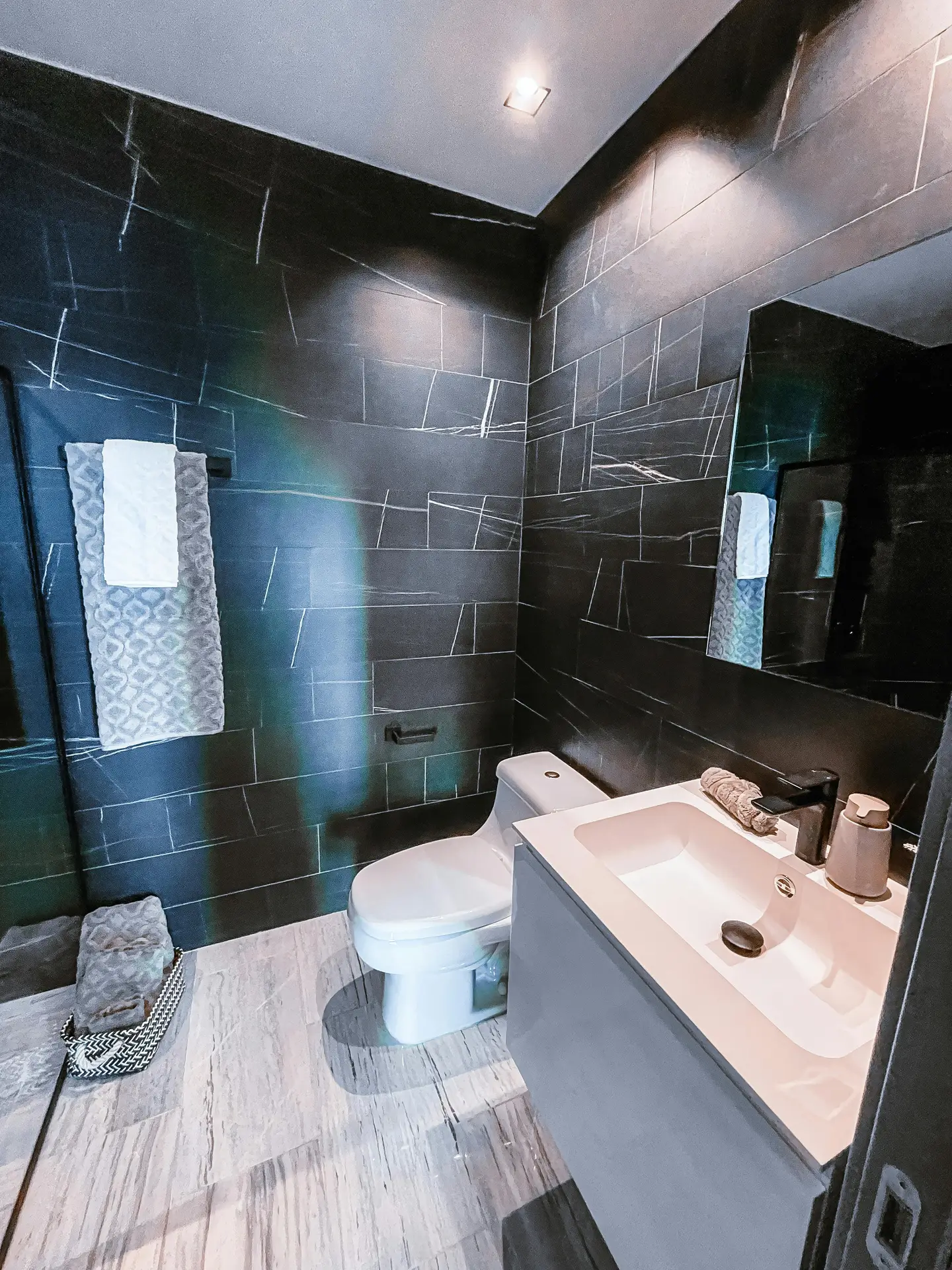 A perspective of a modern powder room showcasing dramatic guest bathroom ideas. The space is defined by matte black tiles walls and a contrasting light floor made of large grey tiles. Centered is a floating oak vanity with a white vessel sink and a black wall-mounted tap, positioned beneath a round black-framed mirror with an LED light above. A sleek, wall-mounted toilet is on the right, with a small framed art piece and a plant on a shelf. A white towel hangs on a black ring.