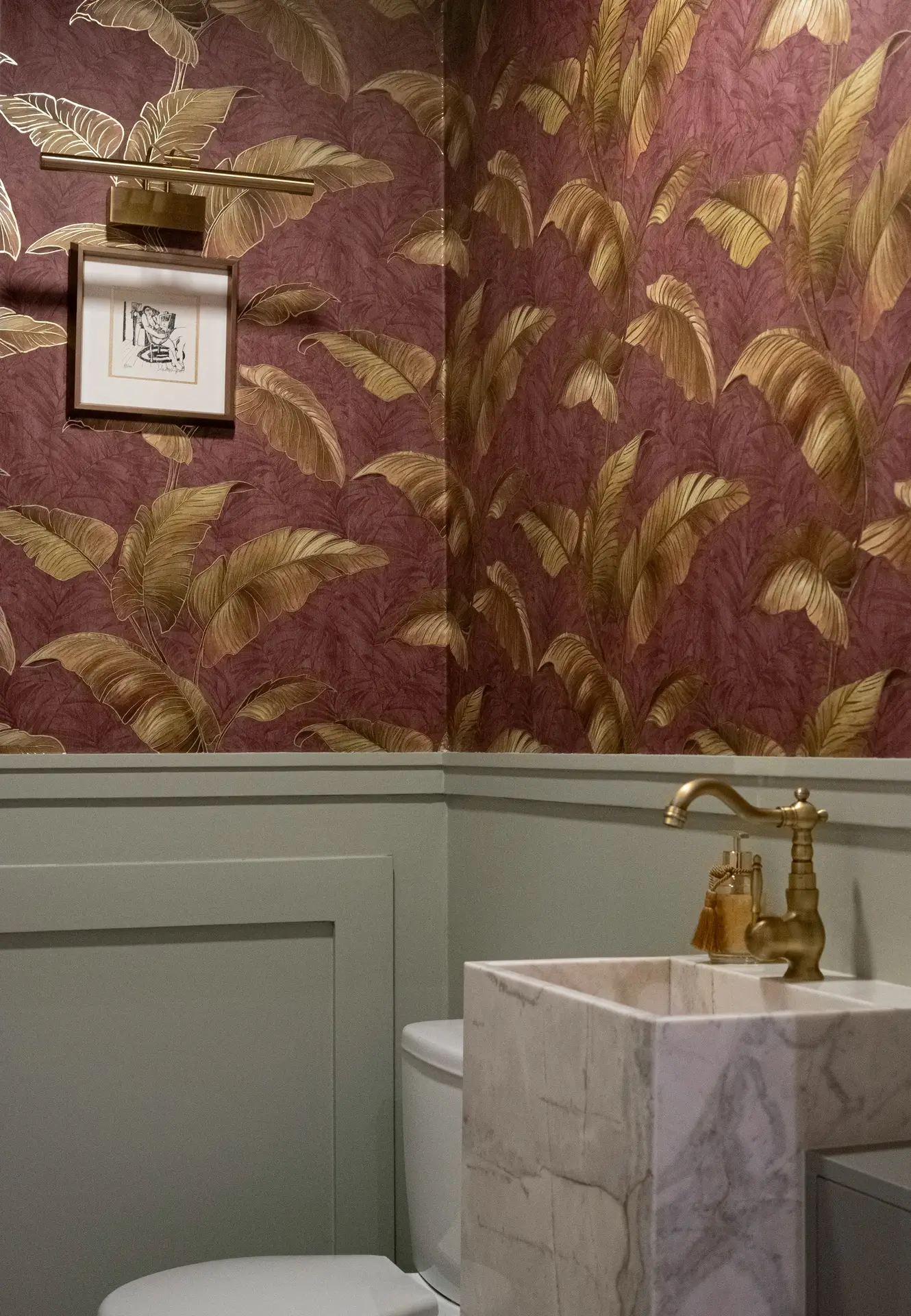 A stylish powder room showcasing unique guest bathroom ideas. The upper walls feature a colorful wallpaper with large gold tropical leaves on a dark burgundy background, contrasting elegantly with the sage green wainscoting below. The space includes a square marble pedestal sink with a vintage brass faucet, a toilet, and a small framed artwork illuminated by a brass picture light.