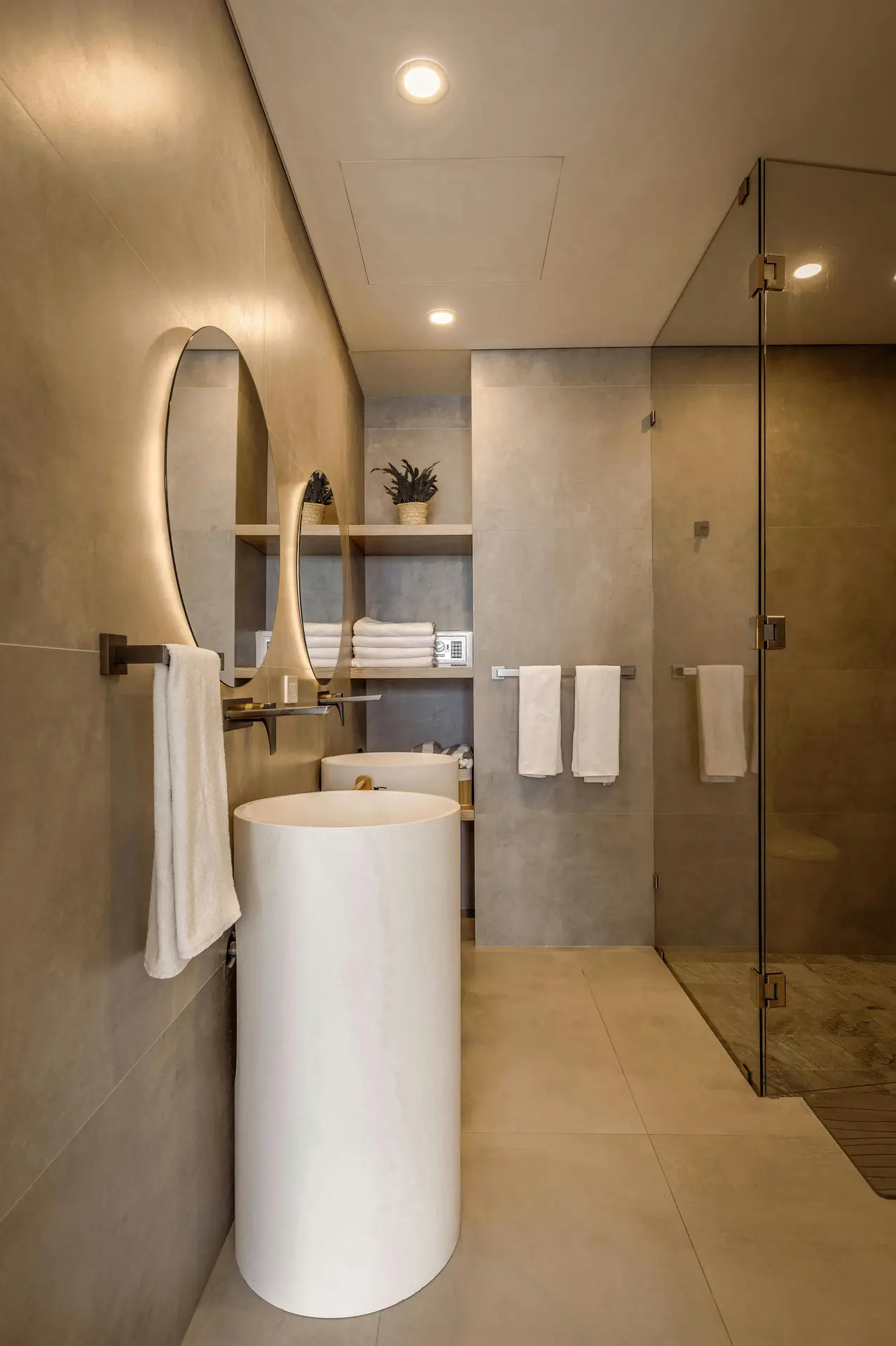A sophisticated, modern bathroom interior highlights minimalist guest bathroom ideas with a focus on geometric forms. Two white cylindrical column sinks stand as sculptural focal points against large-format greige wall tiles. Above the basins, two circular backlit mirrors provide a soft glow, complemented by recessed ceiling spotlights. A sleek glass-enclosed shower is visible on the right, while the back alcove features wooden floating shelves neatly stocked with white towels and a small potted plant, creating an organized and high-end feel.