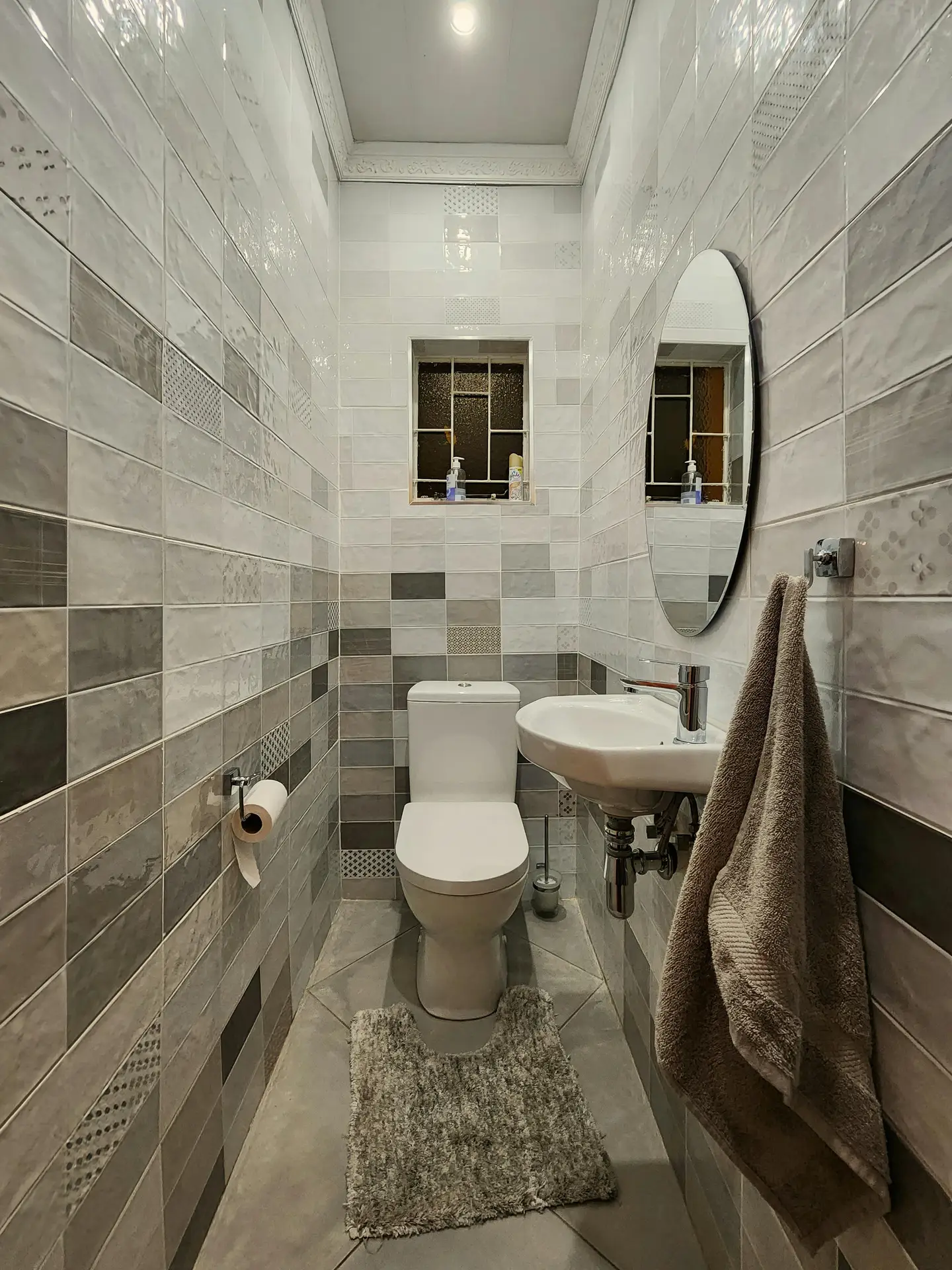 A narrow powder room illustrating practical guest bathroom ideas for tight spaces. The walls are clad in horizontal rectangular tiles in mixed shades, creating a design where almost everything grey is blended with white and subtle textured patterns. At the far end, a white toilet sits beneath a small, deeply set window with a dark frame. On the right, a compact wall-mounted sink with exposed chrome plumbing is positioned under an oval frameless mirror, with a textured towel hanging nearby. A fluffy bath mat rests on the large, square tiled floor.