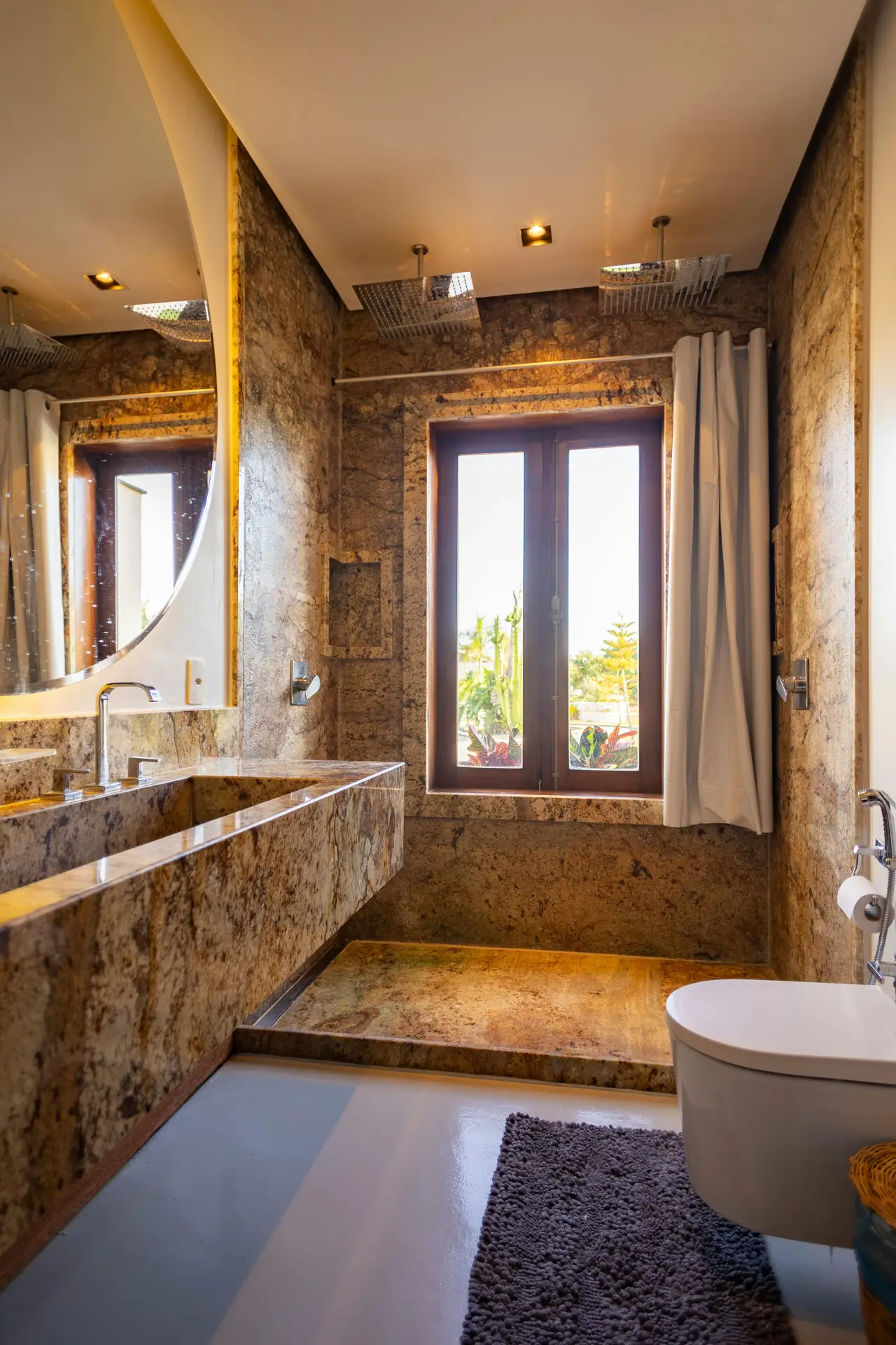 A luxurious bathroom interior offering opulent guest bathroom ideas, dominated by rich brown and gold stone surfaces. This striking granite guest bathroom features a massive floating stone vanity with an integrated sink positioned beneath a large round mirror. The open wet room-style shower at the back includes dual square ceiling-mounted showerheads and a wooden window framing a tropical outdoor view, complete with a side-swept white curtain. A modern white wall-mounted toilet and a textured grey bath mat sit on the smooth, light-colored floor in the foreground.