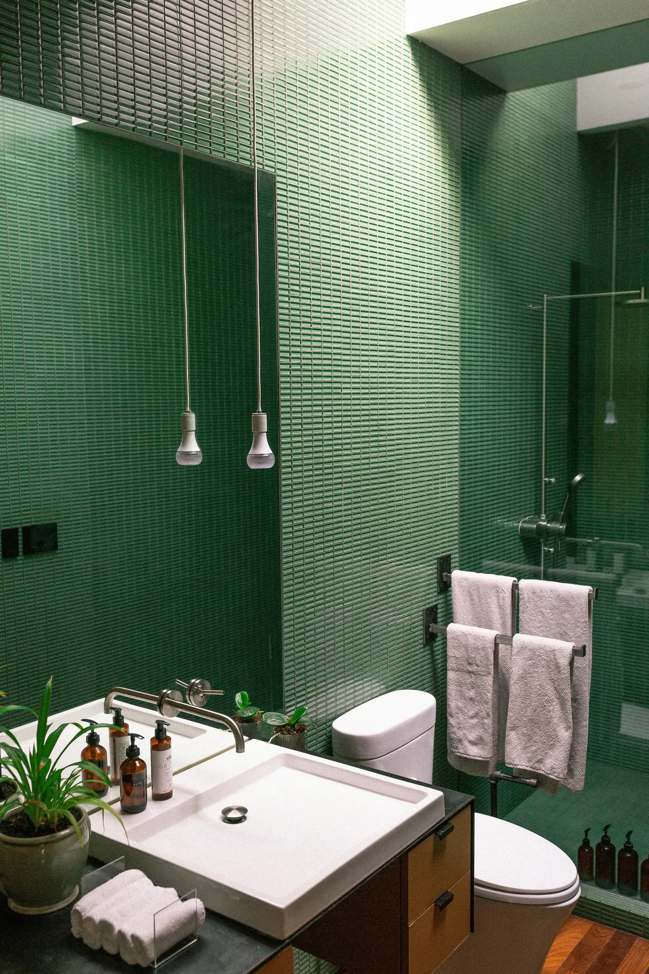 An eye-catching interior packed with guest bathroom ideas, featuring walls entirely enveloped in glossy green small tiles. The space includes a sleek, shallow white square sink on a black and wood vanity, equipped with a wall-mounted silver faucet and amber dispenser bottles. Two minimalist bare-bulb pendants hang in front of a large, frameless mirror. A white toilet, a freestanding metal towel rack holding white towels, and a glass shower enclosure complete the vibrant room above a warm wood floor.