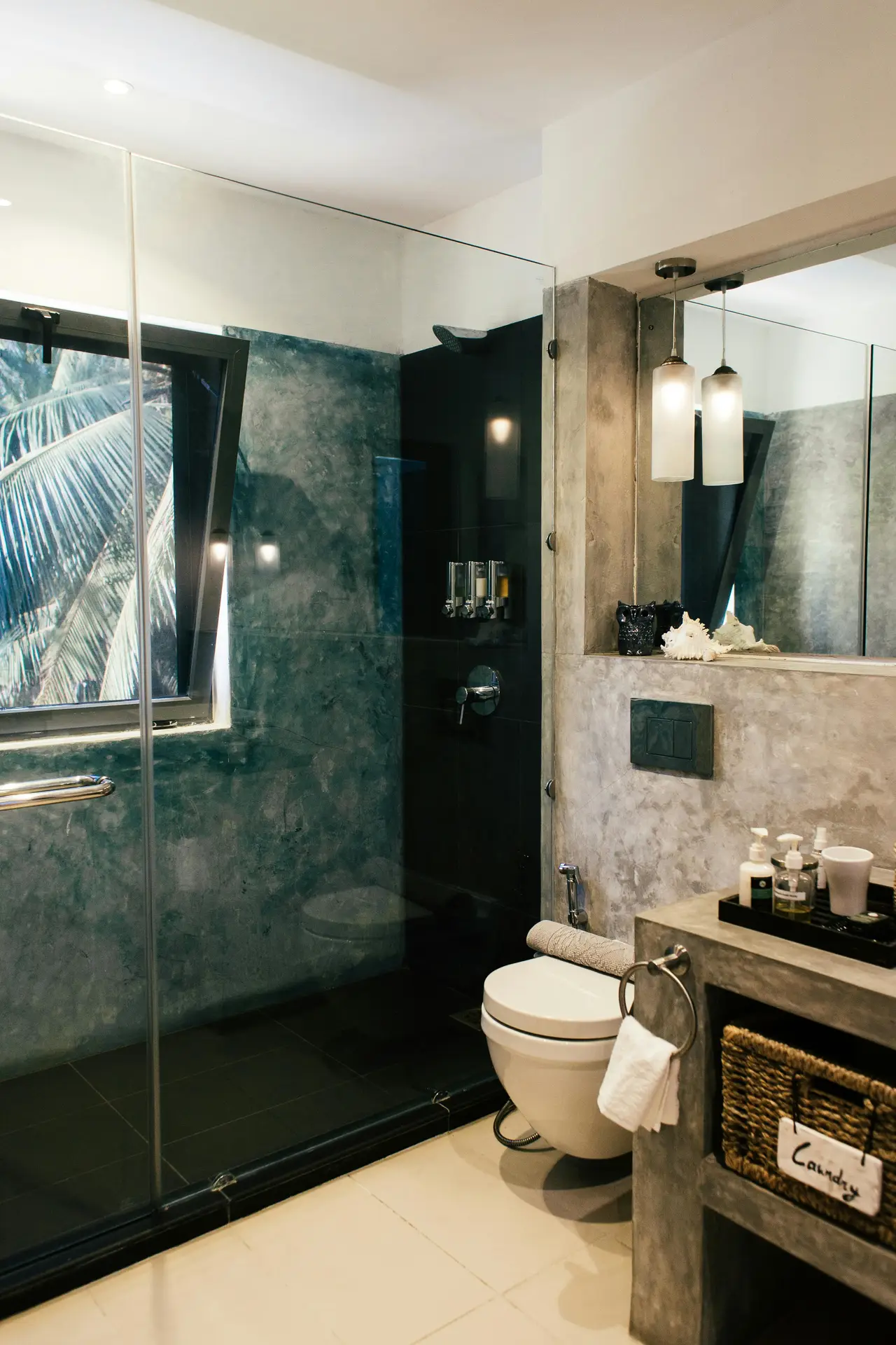 A contemporary guest bathroom featuring a stunning grey stone and black combo is rendered in three distinct perspectives, offering stylish and space-efficient guest bathroom ideas. Large-format grey marble-patterned tiles cover the floor and walls, creating a clean backdrop. The central focus is a black fluted, wall-mounted vanity with an integral circular concrete-grey sink and a tall black faucet. Above it, a large, round mirror is flanked by vertical brass-and-black sconce lights. The other views show a sleek, walk-in glass-enclosed shower on the left and a wall-mounted black toilet on the right. Accents include light wood floating shelves with rolled towels and decorative items, and a potted plant for a touch of life.