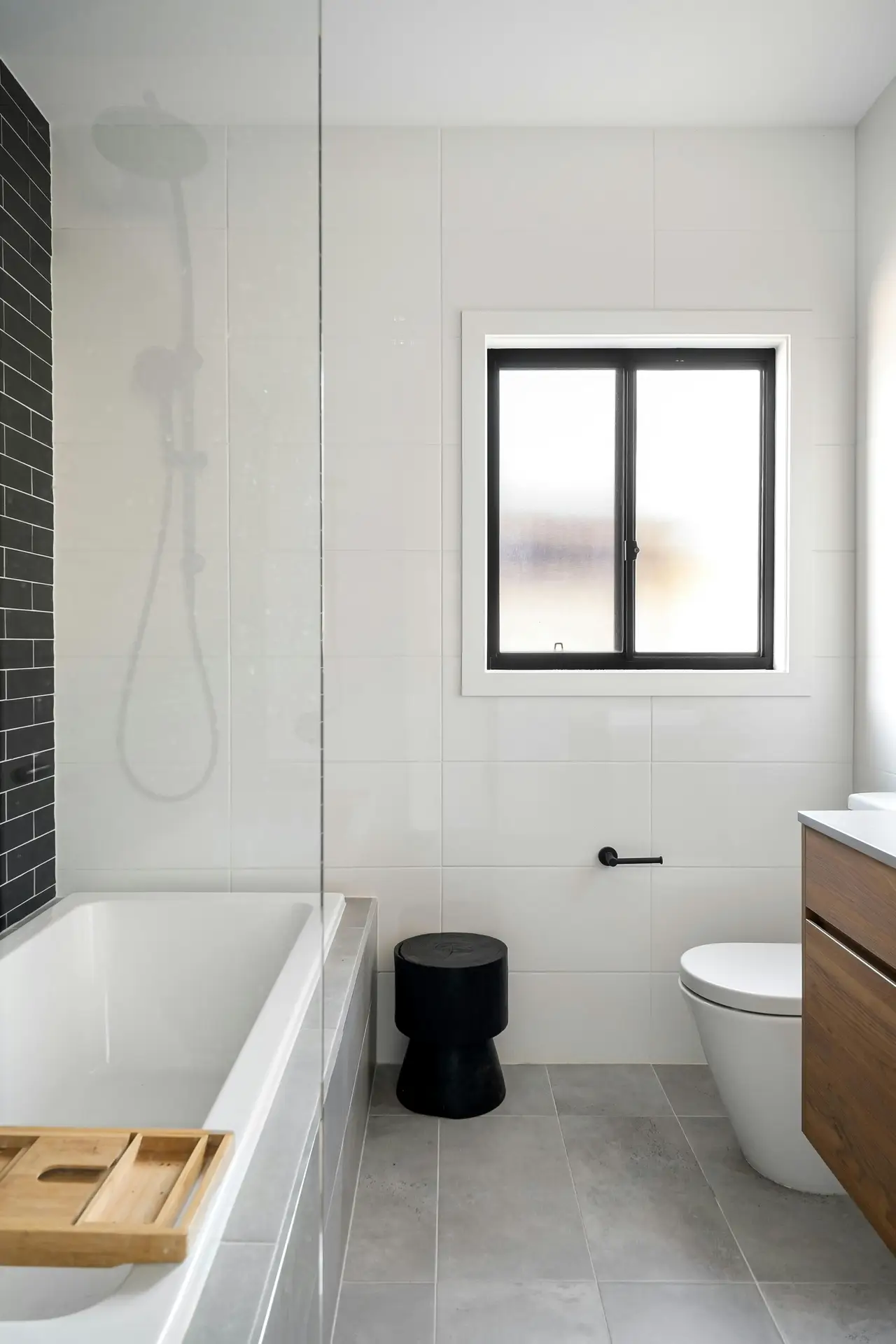 A clean, modern bathroom interior featuring a minimalist color palette of grey, white and black. Large white tiles cover the walls, while the floor is laid with matte grey tiles. The space provides several guest bathroom ideas, including a built-in bathtub on the left with a glass splash screen and a black tiled accent wall behind the shower. A sleek white toilet sits next to a wooden vanity, and a black pedestal stool is placed under a black-framed window. A light wood bath caddy rests on the tub, adding a warm touch to the contemporary design.