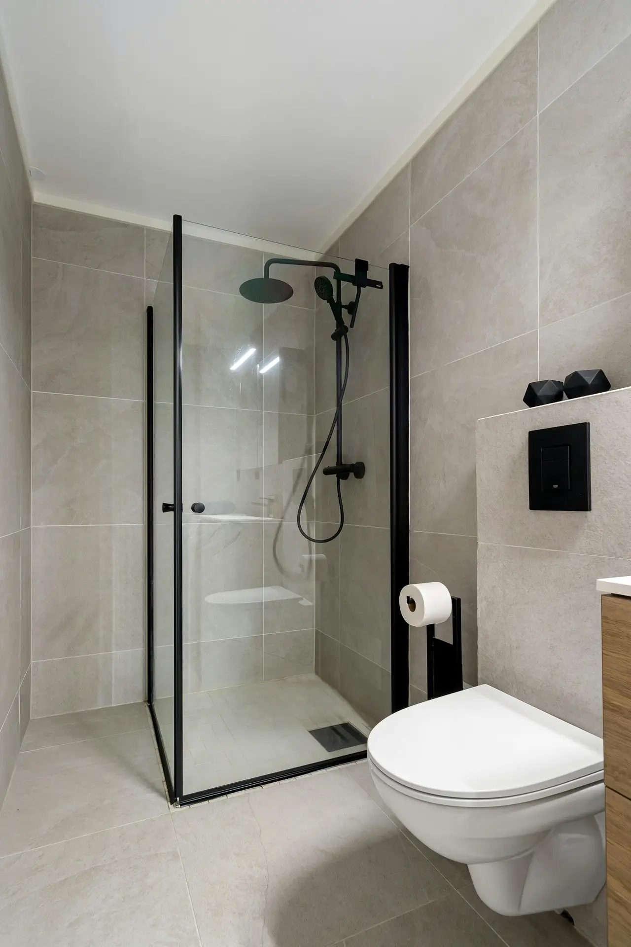 A contemporary interior view showcasing a modern guest bathroom defined by a sleek color palette of light grey with black accents. Large-format, polished grey marble tiles clad the floor and walls, creating a seamless background. The room features a wall-mounted ceramic vanity with an integrated basin and a black industrial-style frame below, paired with matching black square tapware and a tall, thin vertical radiator. Above the sink hangs a large, round mirror with a thin black frame, flanked by two black geometric sconce lights. A glass screen encloses the shower area with a black frame and a rainfall showerhead. On the left, a built-in toilet is positioned next to an oak-topped ledge. Various guest bathroom ideas are incorporated, including floating light wood shelves stocked with neat white towels, toiletries, and small potted plants for a spa-like feel. The ceiling is simple with recessed spotlights.
