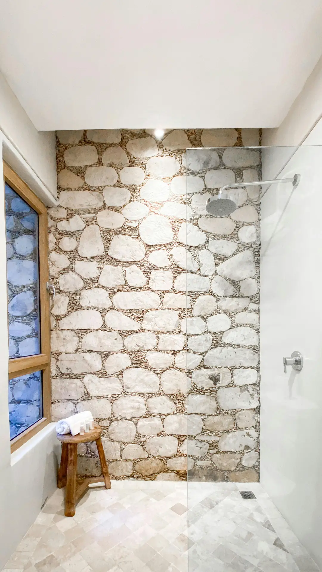 A natural-light filled room displays unique guest bathroom ideas, where a rugged, natural stone wall forms the rugged back and side enclosure for a spacious walk-in shower with a large rain head. A floating, thick rough-edge wood slab vanity with an integrated trough sink sits opposite, illuminated by three stylish pendant lights. The floor features smooth, large-format grey tiles. Above the natural stone wall, a linear glass window allows additional light and views of greenery. The ceiling is simple with exposed wood beams. A minimalist chrome and wood bench and a few woven items complete the space.