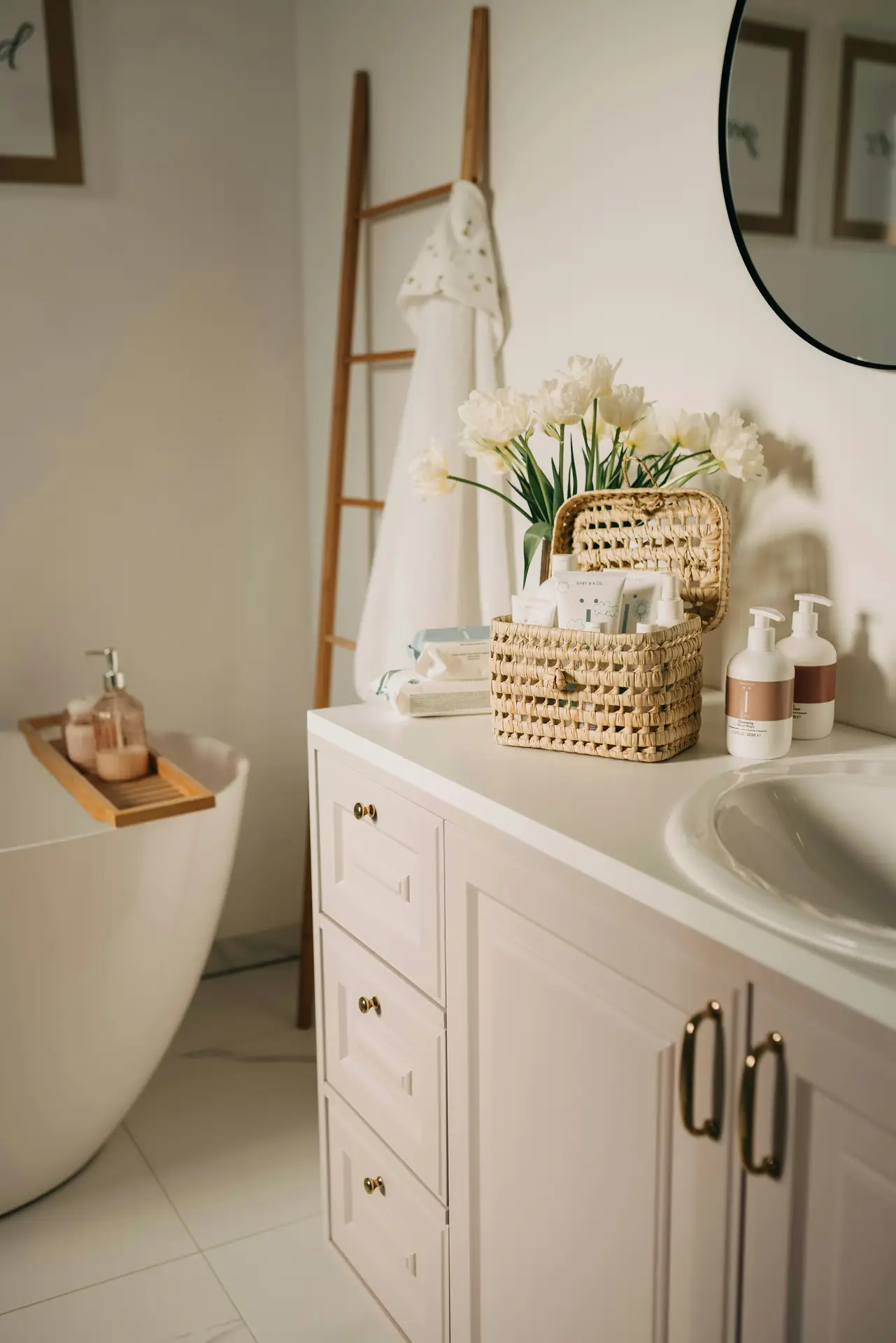 A welcoming bright bathroom scene showing how to perfectly prepare your guest bathroom for your guests. On a white vanity with brass hardware, a small woven basket is filled with complimentary toiletries alongside full-sized lotion bottles and a vase of fresh white tulips, presenting thoughtful guest bathroom ideas. In the background, a large white freestanding tub features a wooden bath tray, and a wooden blanket ladder leans against the wall holding a white towel.