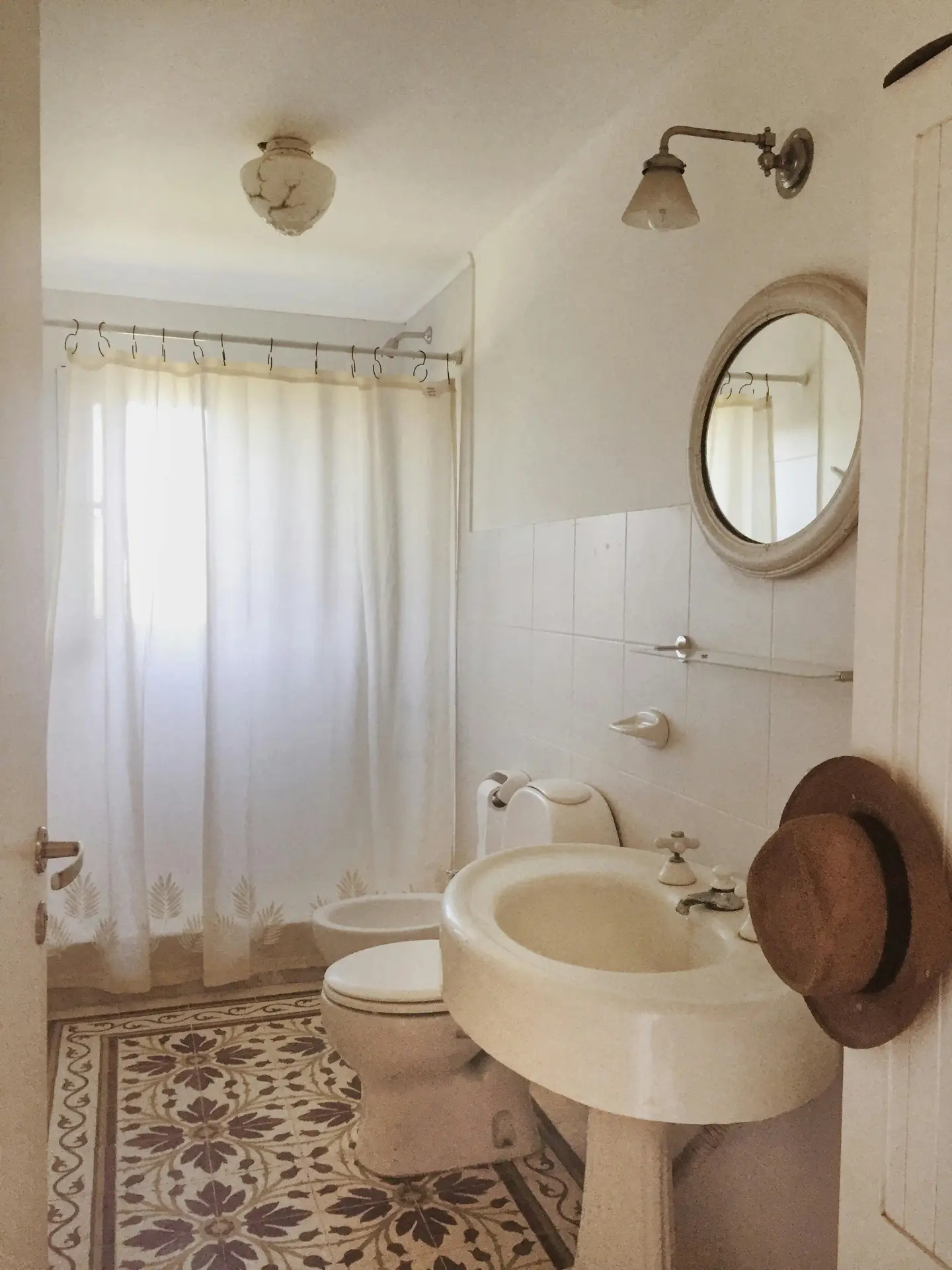 A bright, nostalgic space filled with character, this vintage guest bathroom features intricate floral-patterned floor tiles in shades of brown and cream. The classic layout provides various guest bathroom ideas, such as a large cream pedestal sink with traditional cross-handle faucets, a matching toilet, and a bidet. A round, ornate mirror is mounted on the white tiled wall beneath a swan-neck sconce lamp, while a white shower curtain with a subtle botanical border hangs at the back. A straw hat hangs on the door frame, adding to the cozy, lived-in feel.