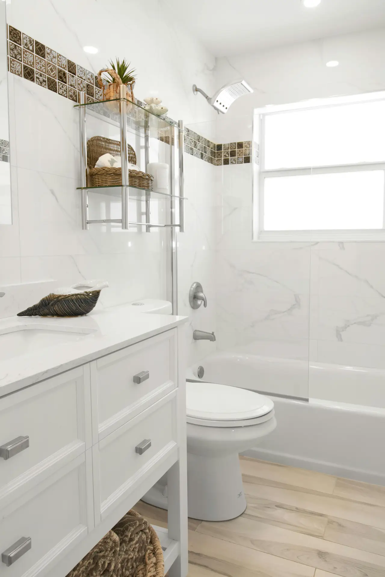 A bright and airy bathroom showcasing refreshing guest bathroom ideas. The space features a clean white and wood combo, highlighting a white vanity with silver hardware, a matching toilet, and a bathtub enclosed by a glass screen. The walls are covered in white marble-look tiles accented by a horizontal band of brown patterned mosaic tiles. Light wood-look plank flooring adds warmth to the room. Above the toilet, a chrome and glass shelving unit holds woven baskets, rolled towels, and a small potted plant, while a large decorative seashell rests on the sink counter near a bright window.