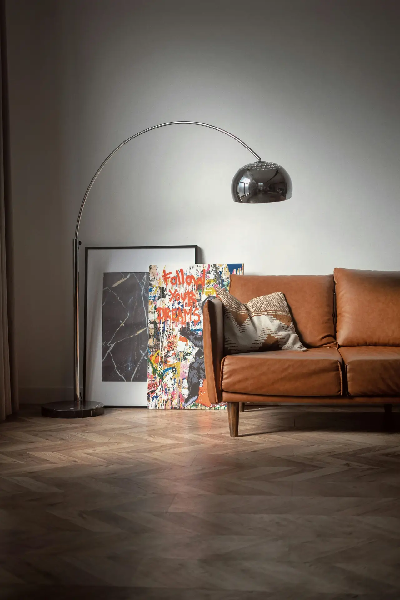 A sleek arc floor lamp with a polished chrome finish and a large dome-shaped metal shade curves elegantly over a tan leather sofa. The lamp stands on a dark marble base next to vibrant modern artwork, creating a sophisticated example of mid century modern lighting in a living room with herringbone wood floors.