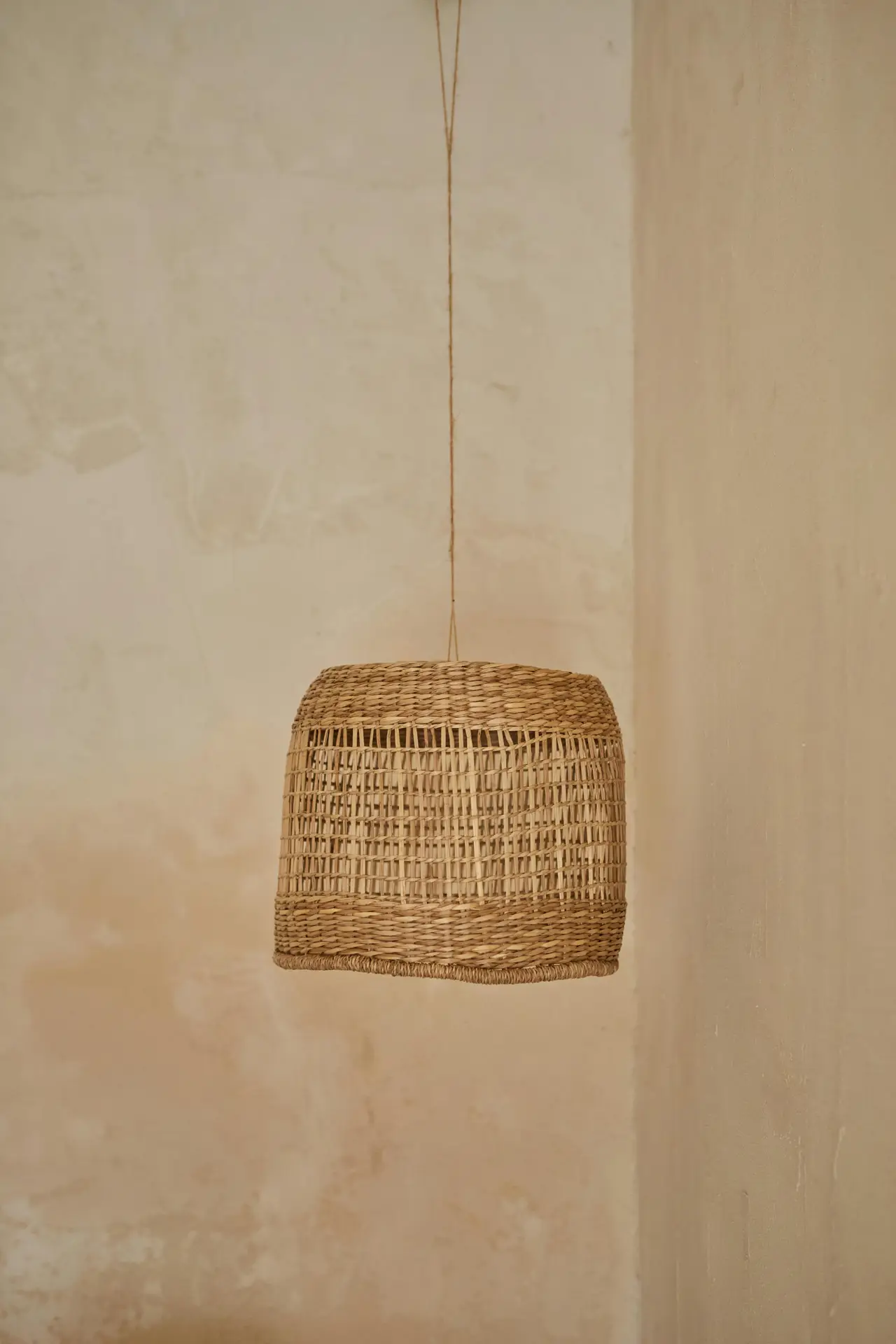 A rustic boho-style woven pendant light made of natural seagrass or rattan hangs from a simple cord against a textured, sandy-colored wall. The intricate basket-weave design adds a warm, organic touch to the space, beautifully bridging the gap between natural textures and mid century modern lighting principles.