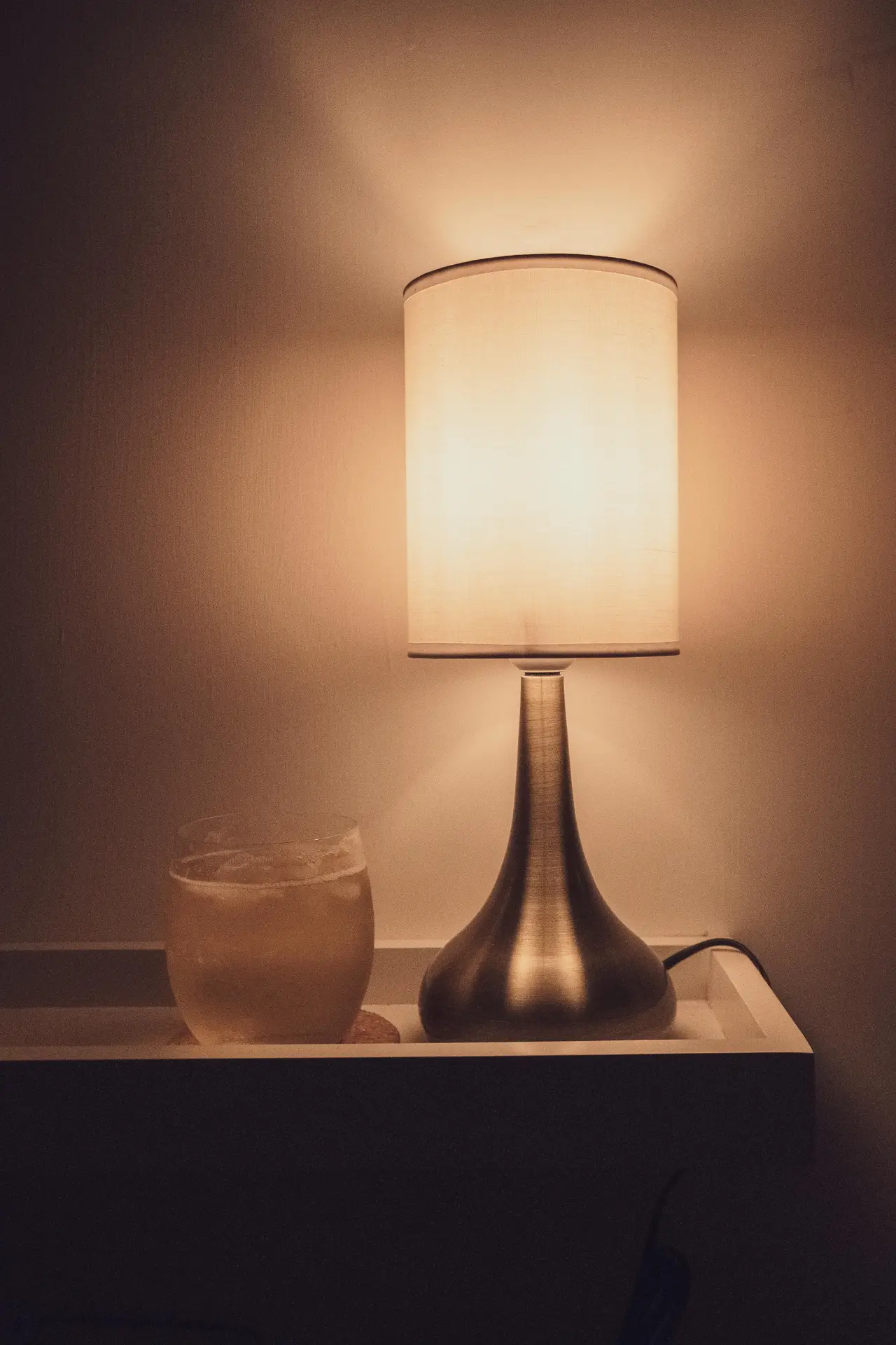 A glowing cylinder shade table lamp with a sleek, brushed metallic teardrop base sits on a white shelf. The warm light creates a cozy ambiance in the dark room, illuminating a glass of iced tea nearby and showcasing a classic example of mid century modern lighting.