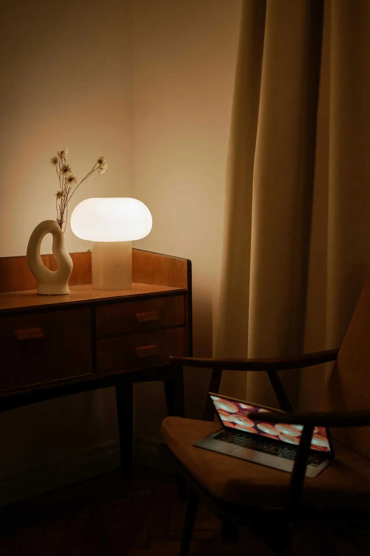 A glowing donut-style lamp with a smooth, rounded white glass shade sits on a vintage wooden sideboard, casting a warm light through the room. Next to it stands a sculptural cream-colored vase, while an open laptop rests on a classic armchair in the foreground, perfectly capturing the cozy atmosphere of mid century modern lighting.