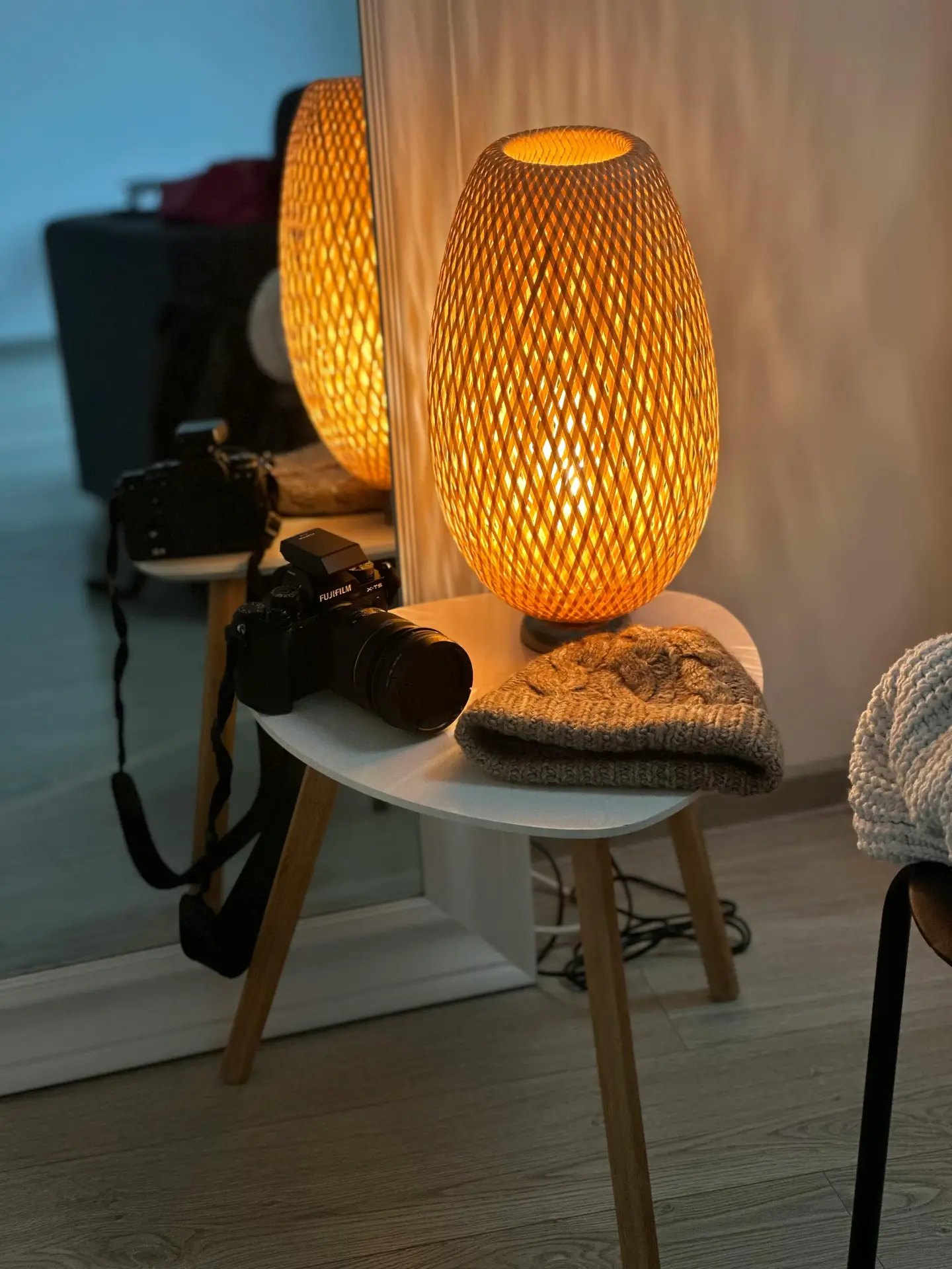 An egg-shaped bamboo lamp with an intricate woven lattice design sits on a white tripod side table, casting a warm, patterned glow against the wall. A Fujifilm camera and a brown knit beanie are placed next to the lamp, creating an inviting corner that highlights the organic textures often found in mid century modern lighting.