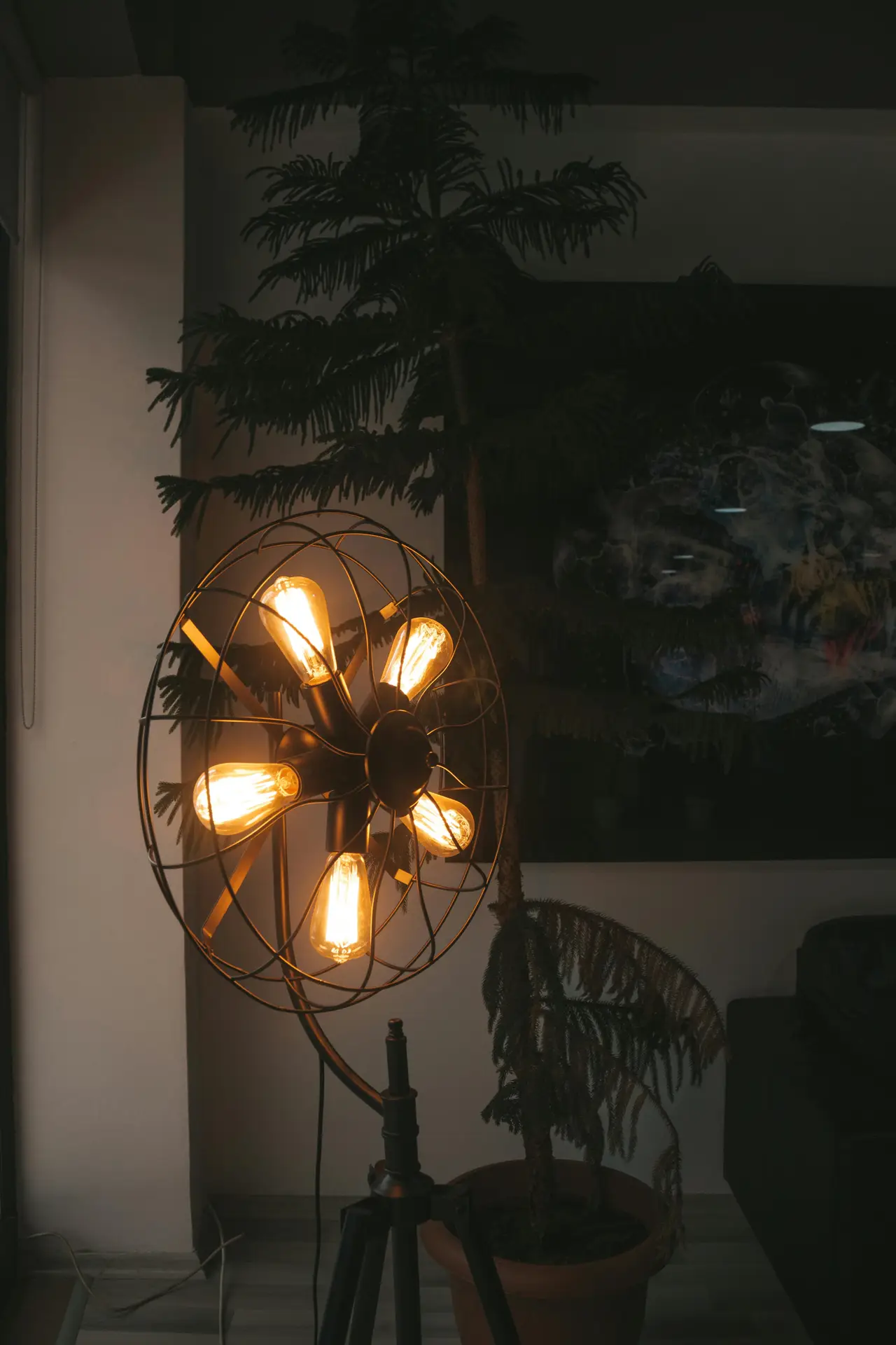 A unique fan-style floor lamp with five glowing Edison bulbs arranged in a circular metal cage, resembling a vintage industrial fan. The warm light creates a cozy atmosphere in a dark room next to a potted Norfolk Island pine, serving as a standout piece of mid century modern lighting.