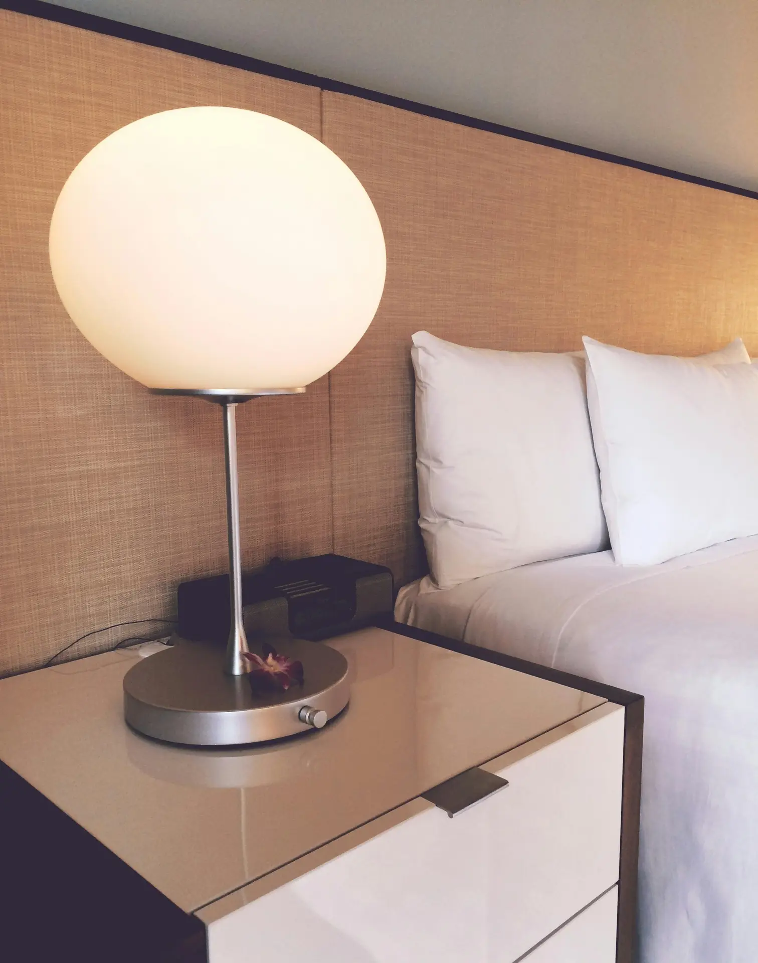 A large, frosted globe table lamp with a sleek brushed-metal base and a slender stem sits on a modern bedside table. The soft, diffused light illuminates a neutral-toned headboard and crisp white bedding, perfectly illustrating a sophisticated piece of mid century modern lighting.