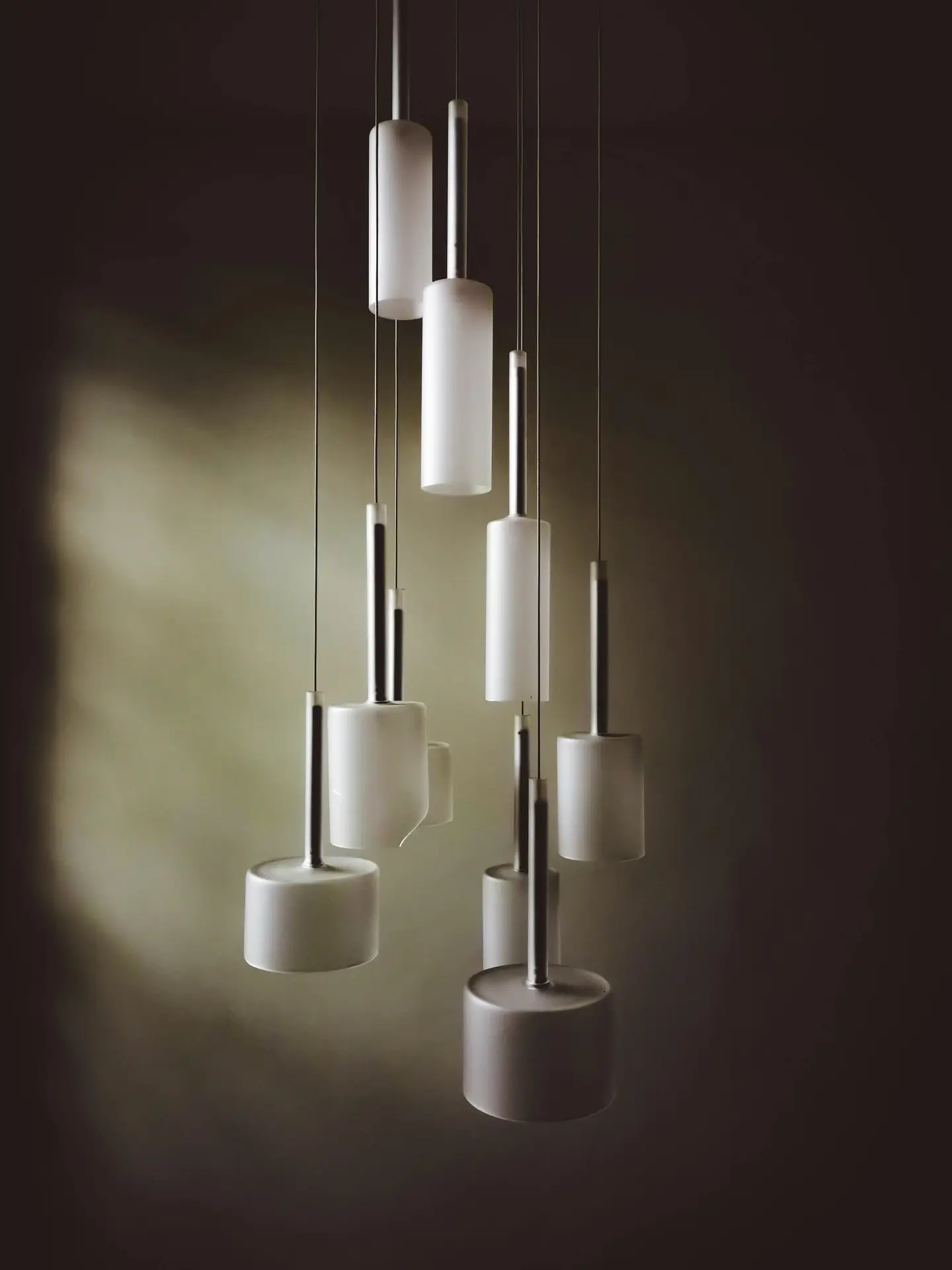 A dramatic multi-light cluster pendant featuring numerous frosted white cylindrical shades hanging at varying lengths. The minimalist, staggered arrangement creates a sculptural focal point against a dark, atmospheric background, showcasing a bold expression of mid century modern lighting.