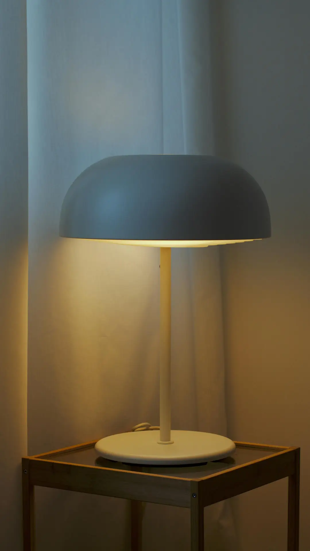 A minimalist mushroom table lamp with a smooth, domed grey shade and a slender white stem stands on a glass-topped wooden side table. The lamp emits a warm, ambient glow against a soft white curtain, serving as a clean and functional example of mid century modern lighting.